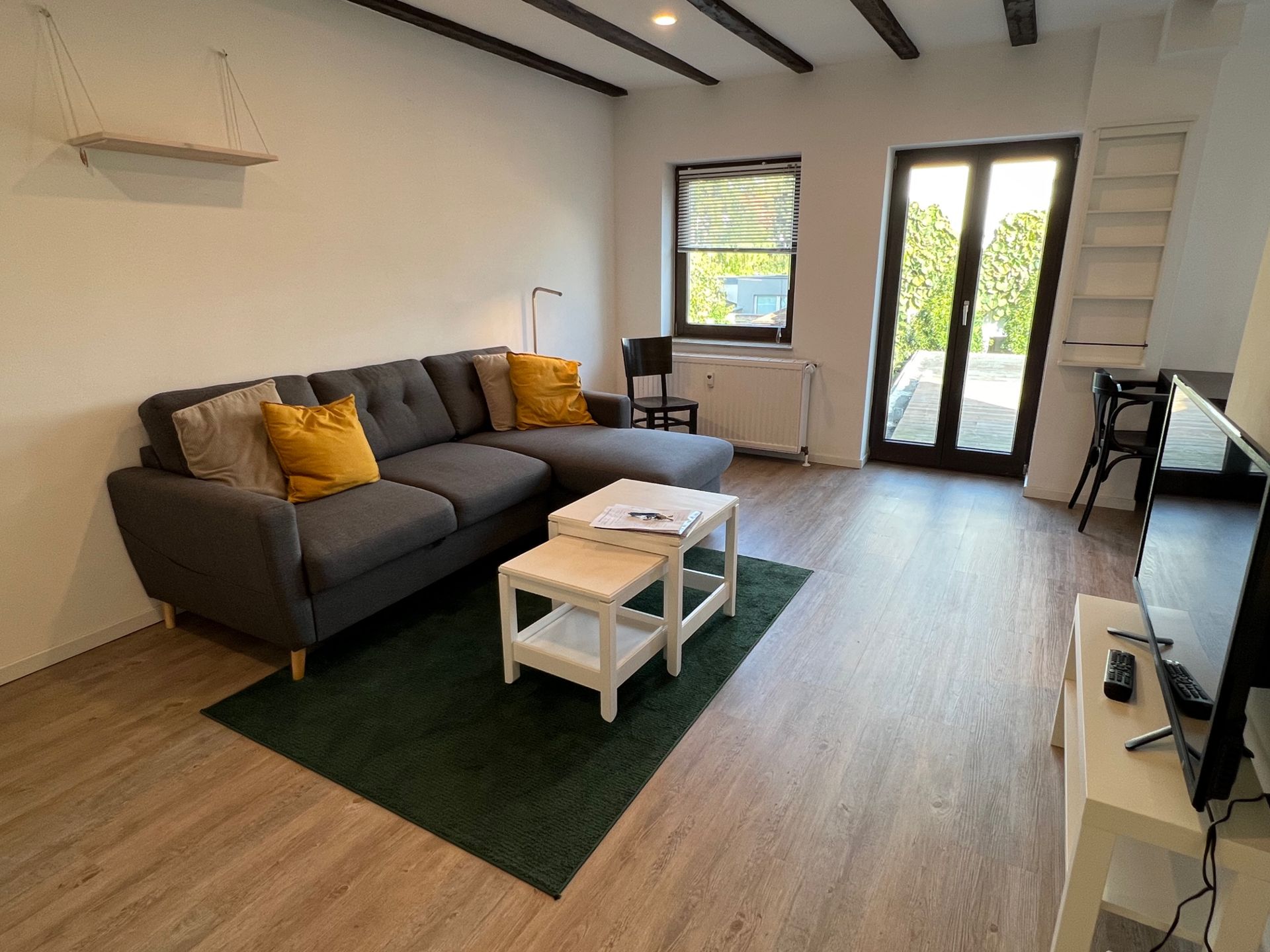 Wohnzimmer mit grauem Sofa, gelben Kissen, Couchtisch, Fernseher und Glastüren zum Garten.