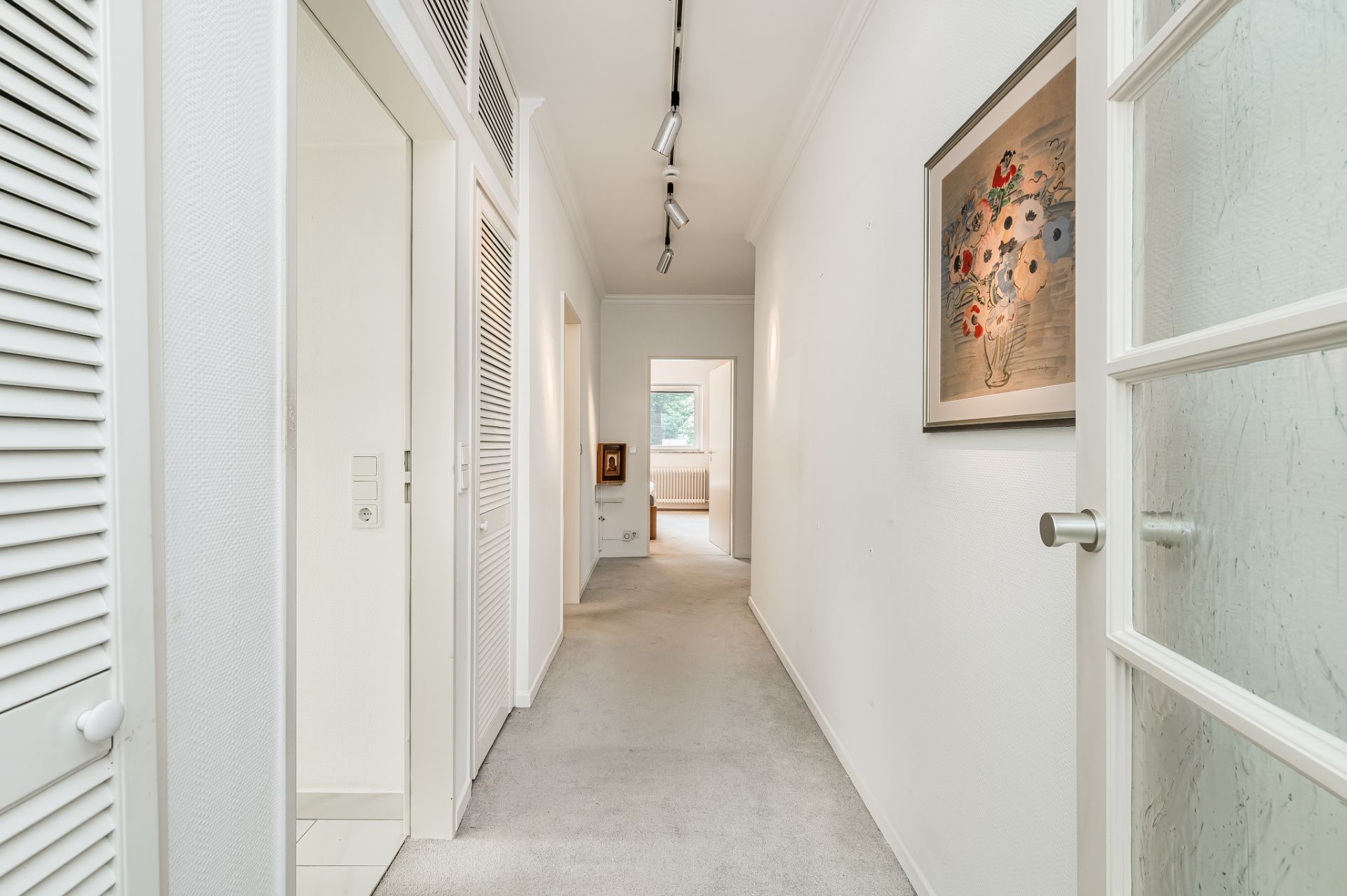 Heller Flur mit Teppichboden, weißen Wänden, Einbauschränken und einem Blumenbild an der Wand.