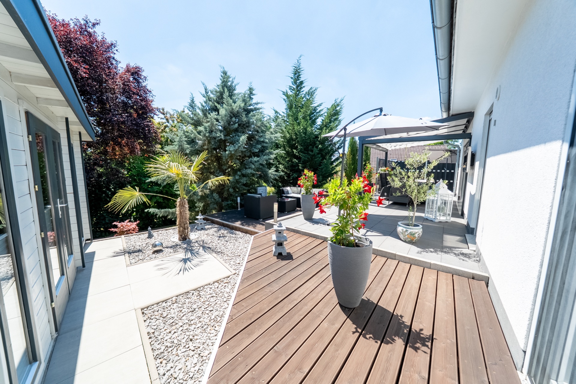 Moderne Terrasse mit Holz- und Steinboden, Pflanzen und Gartenmöbeln, umgeben von Bäumen.