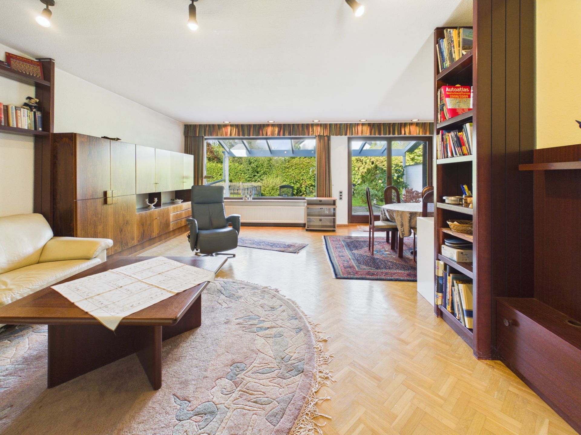 Wohnzimmer mit Bücherregal, Sofa, Sessel, großem Fenster und Esstisch, Blick auf Garten.