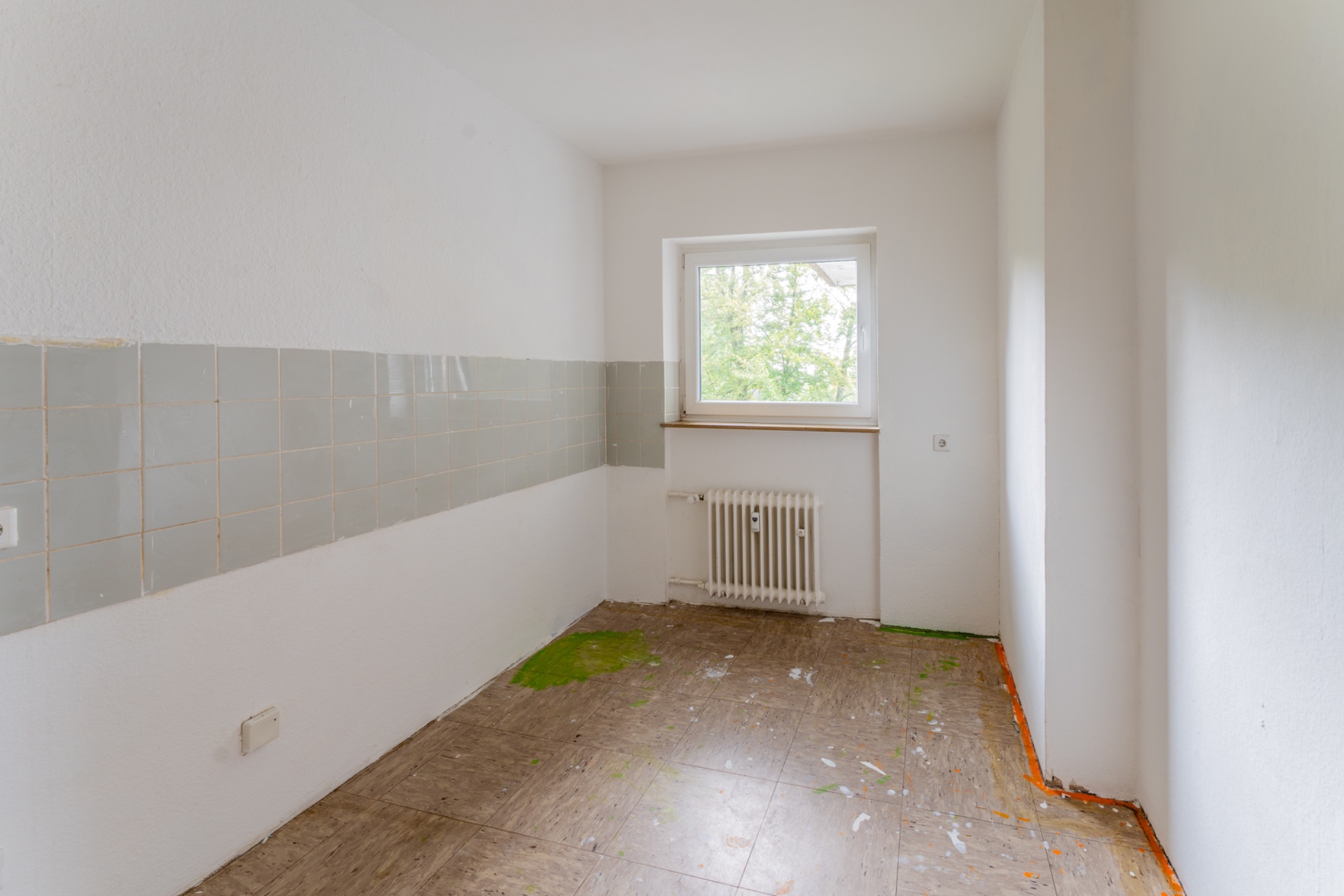 Leerer Raum mit Fliesen an der Wand, Fenster und Heizkörper, Boden teilweise mit Farbflecken.