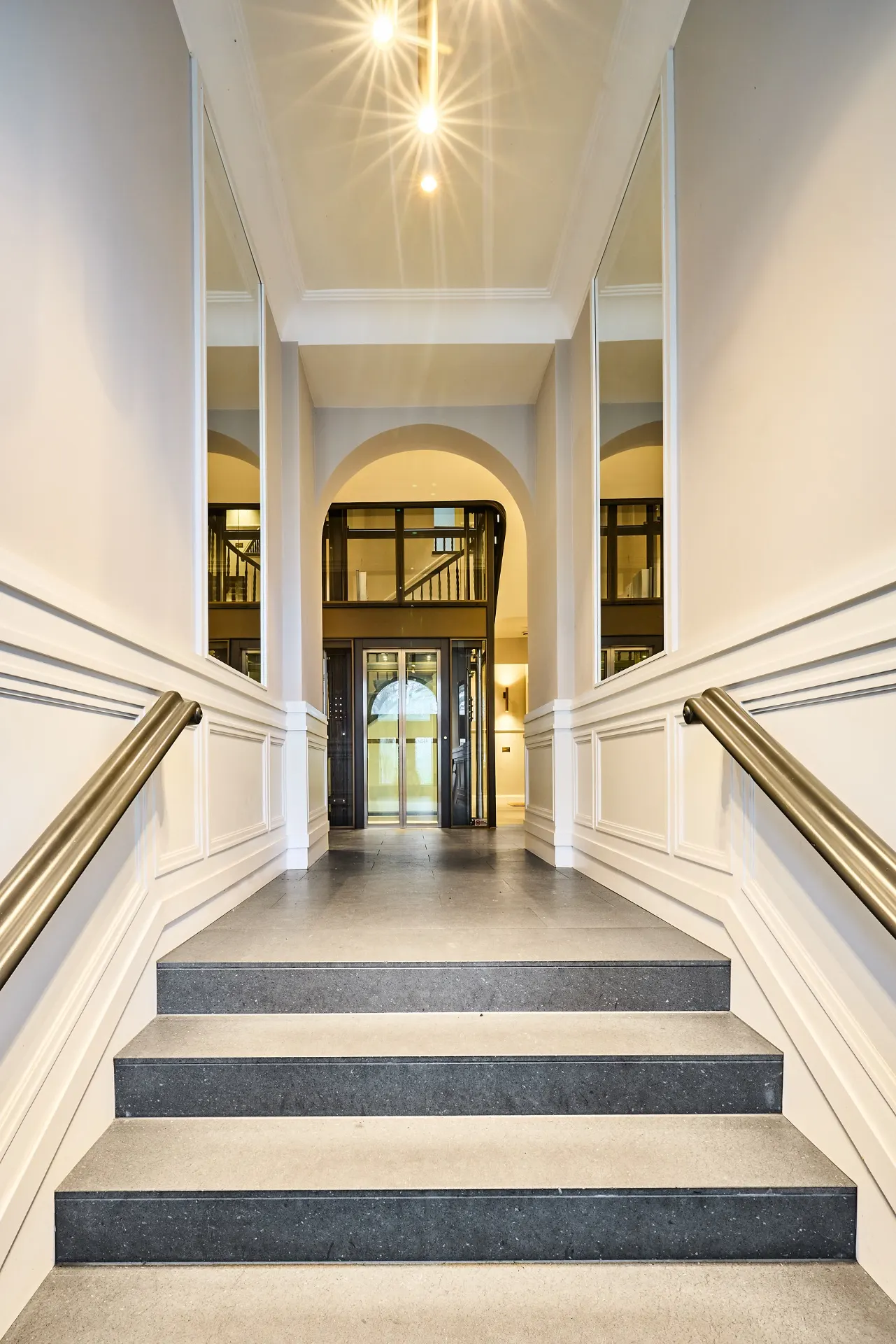 Heller Hausflur mit Treppe, Handläufen und einem Aufzug am Ende.