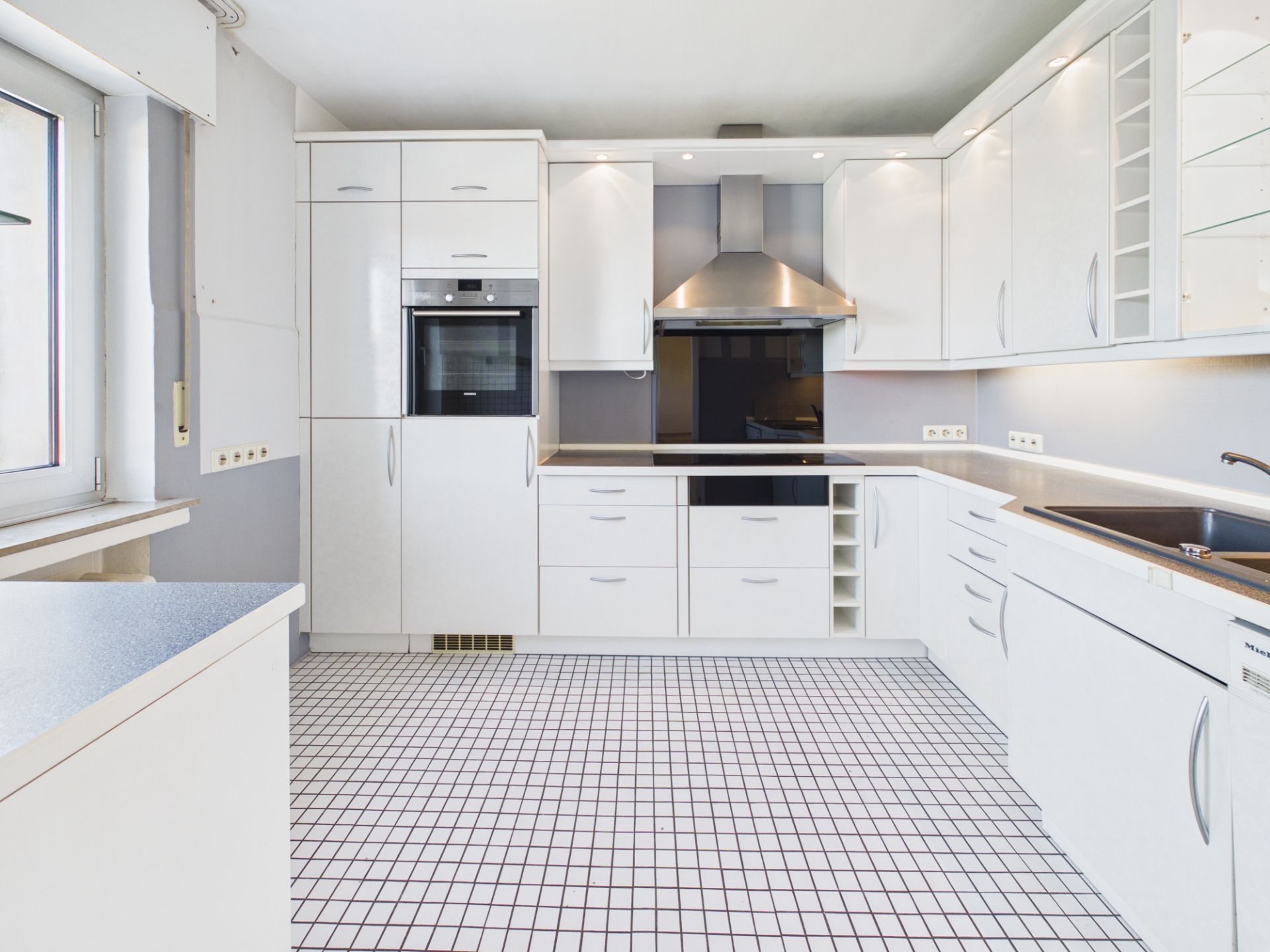 Moderne weiße Einbauküche mit Fenster, Herd, Backofen und Fliesengitterboden.