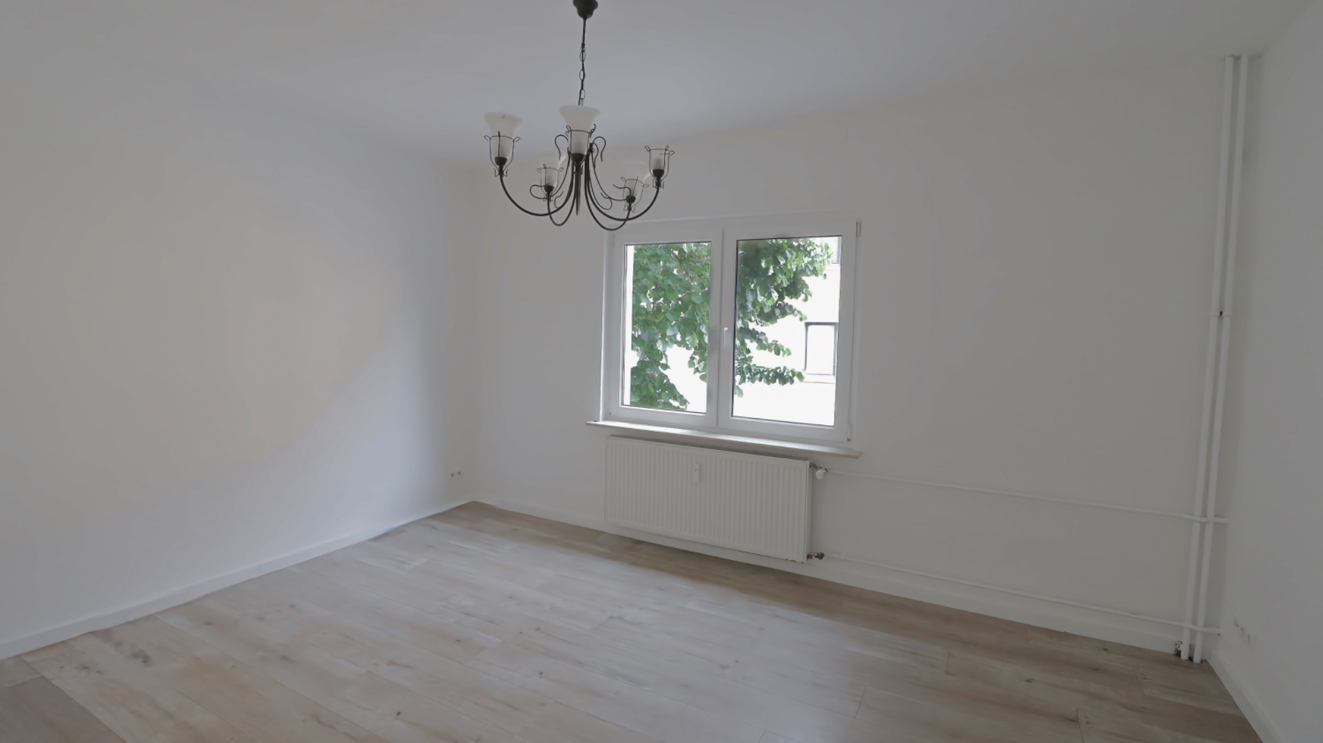 Leeres Zimmer mit Holzfußboden, Fenster, Heizung und Kronleuchter.