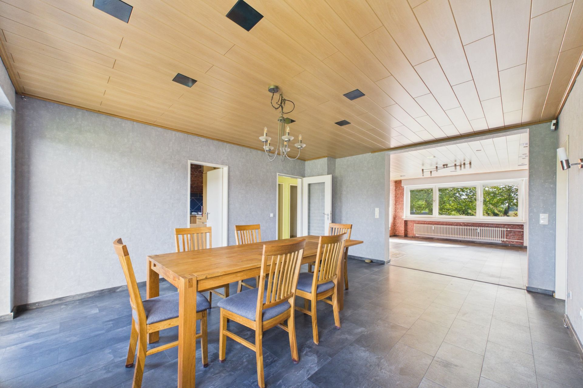 Esszimmer mit Holztisch und sechs Stühlen, angrenzender Raum mit großen Fenstern und grauem Boden.