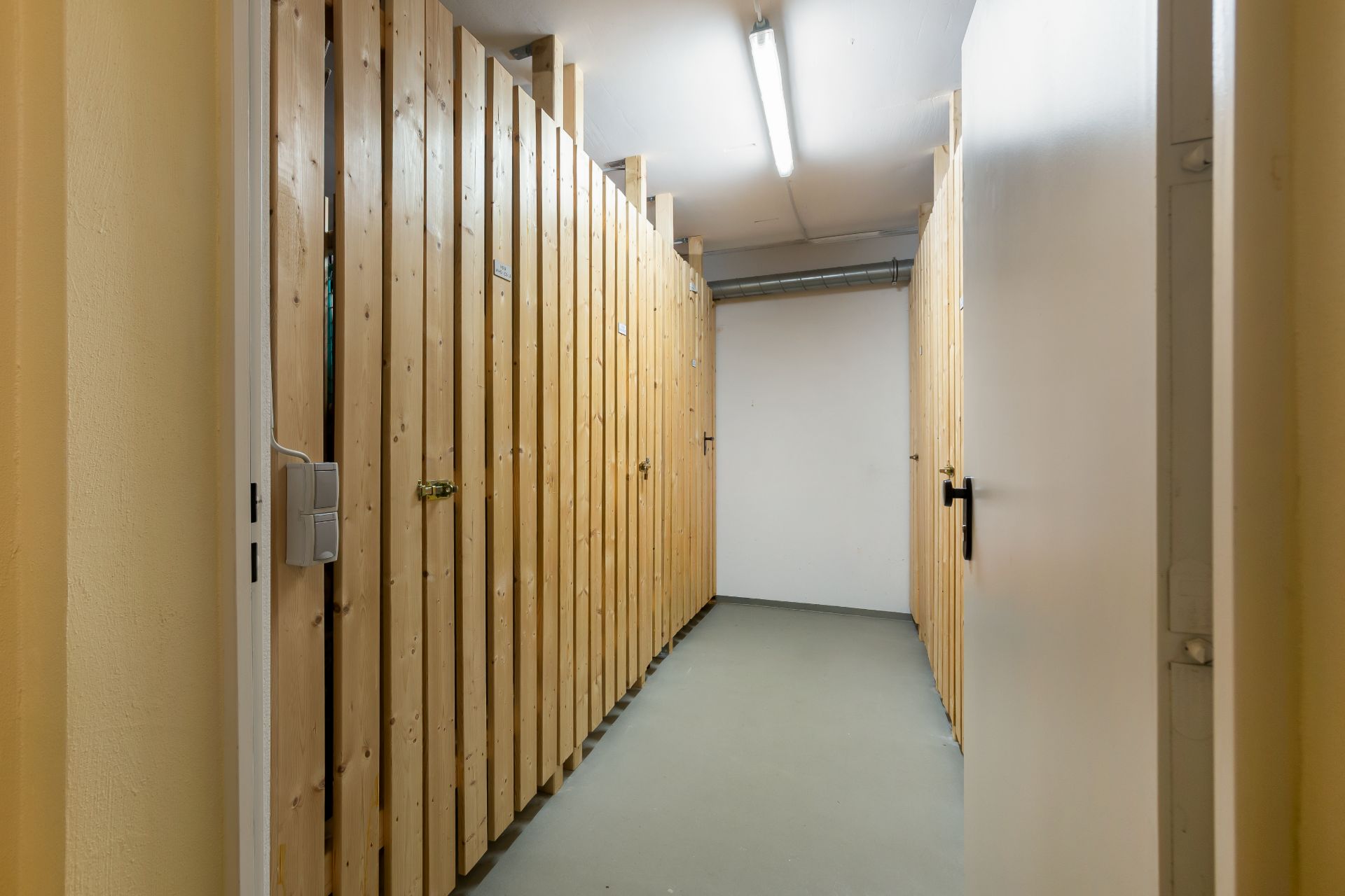 Gang mit Holztüren zu Kellerabteilen in einem Wohnhaus, beleuchtet von einer Deckenlampe.