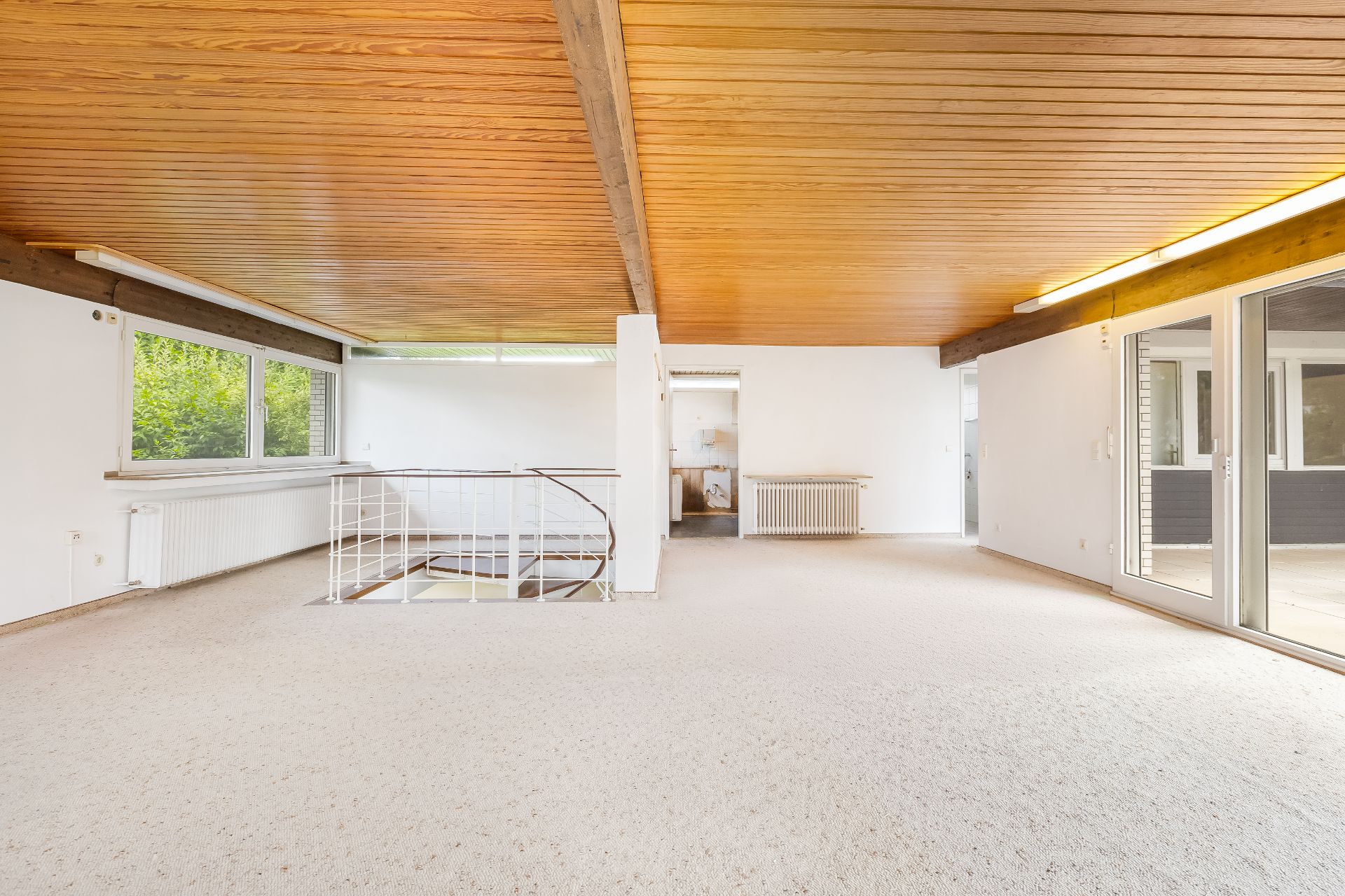 Heller Wohnraum mit Holzdecke, großen Fenstern, Teppichboden und Treppe nach unten.