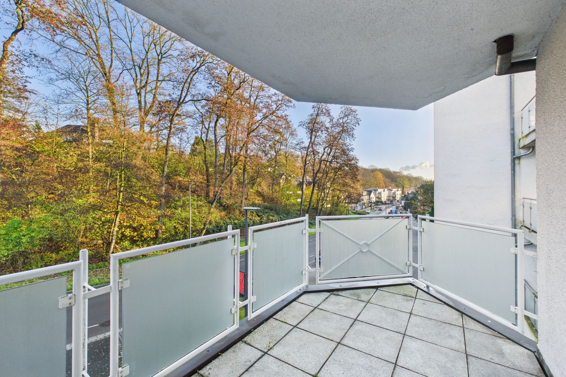 Balkon mit Fliesenboden und Glasgeländer, Blick auf Bäume und eine ruhige Straße.