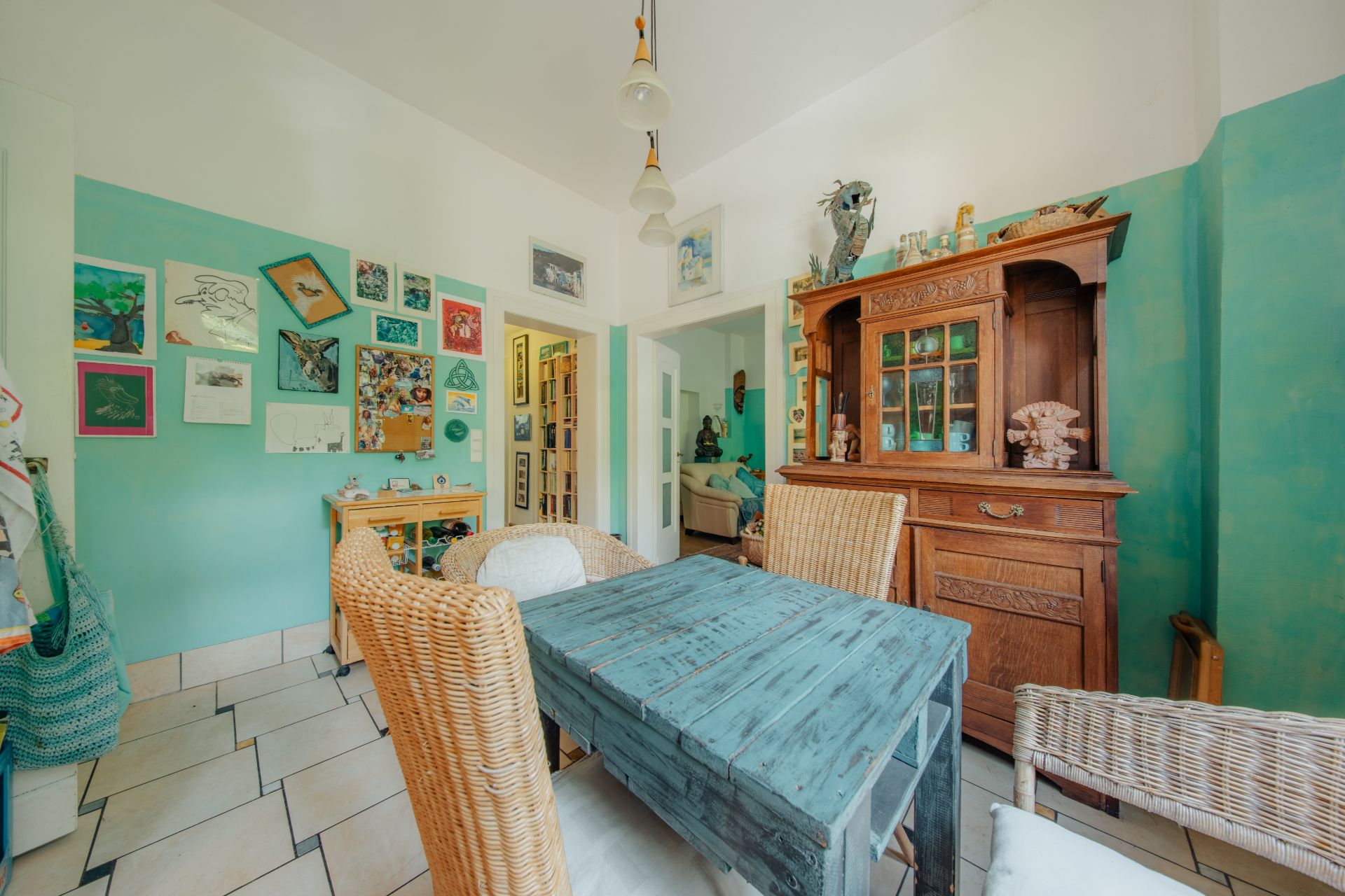 Esszimmer mit Holztisch, Korbstühlen, Holzschrank und Bildern an einer grün gestrichenen Wand.