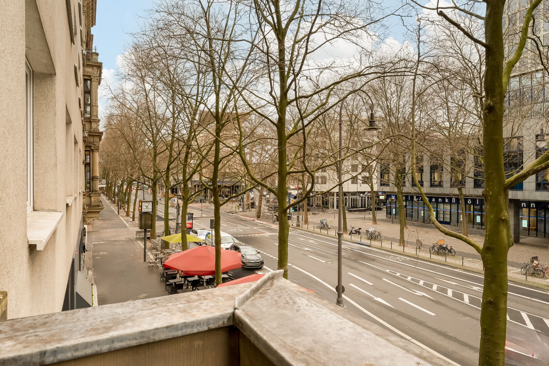 Blick von einem Balkon auf eine Straße mit Bäumen, Tischen mit roten Schirmen und Gebäuden.