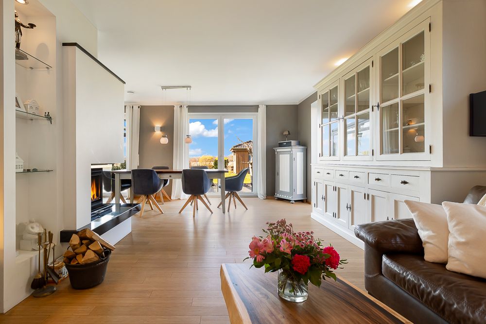 Helles Wohnzimmer mit Kamin, Esstisch, Vitrine und Blumen auf einem Holztisch.
