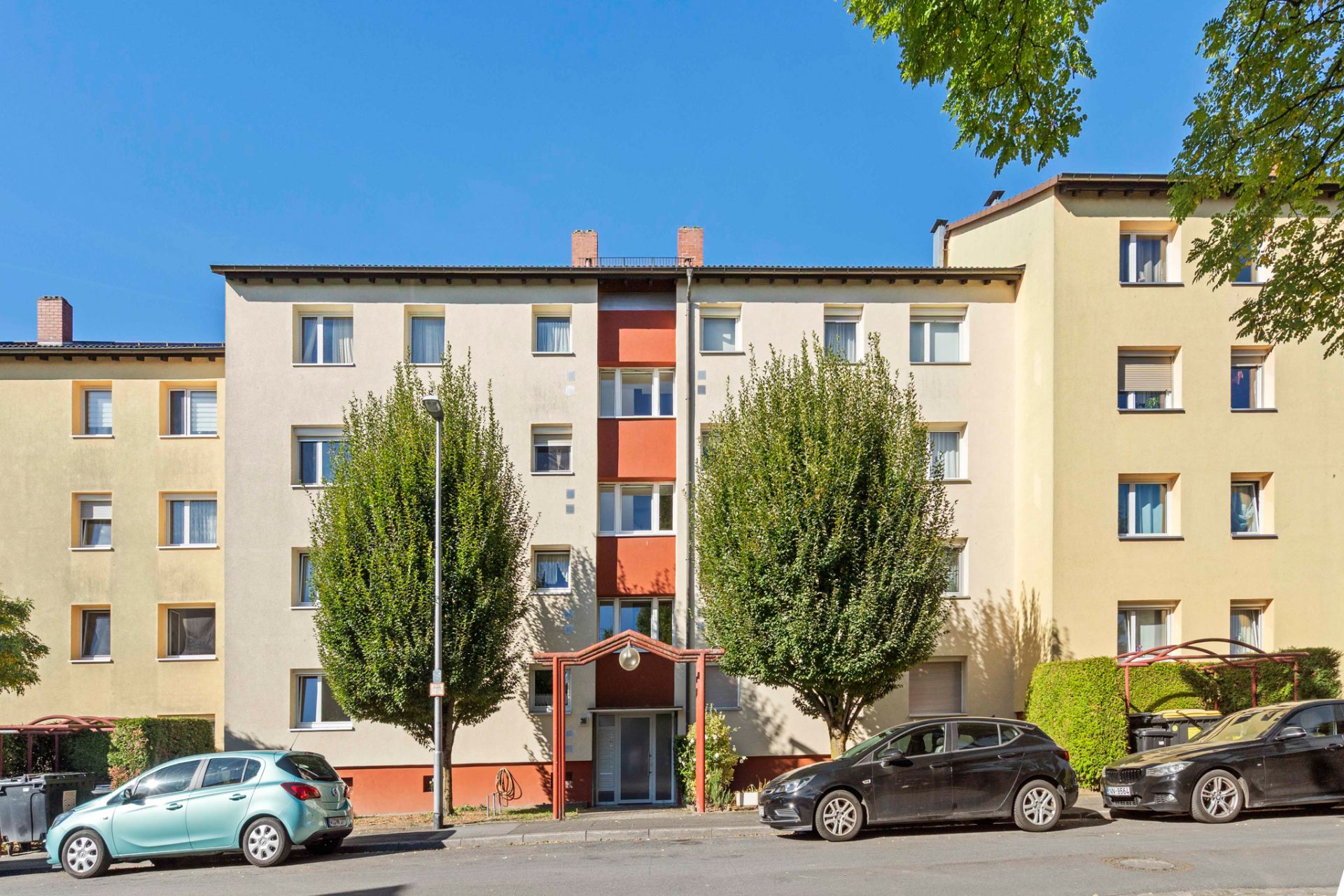 Mehrfamilienhaus mit vier Etagen, Bäumen davor und geparkten Autos an der Straße.