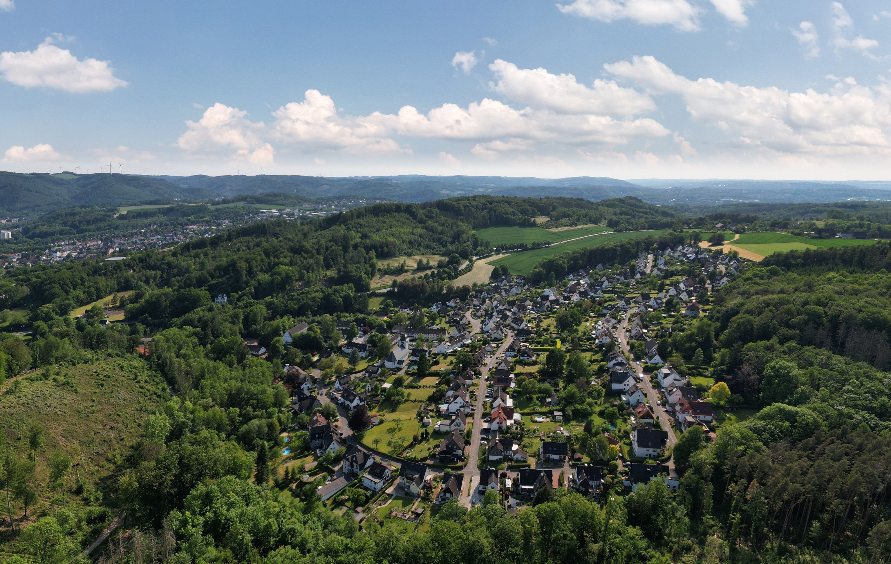 Luftaufnahme einer Wohnsiedlung umgeben von Wäldern und Feldern bei klarem Himmel.