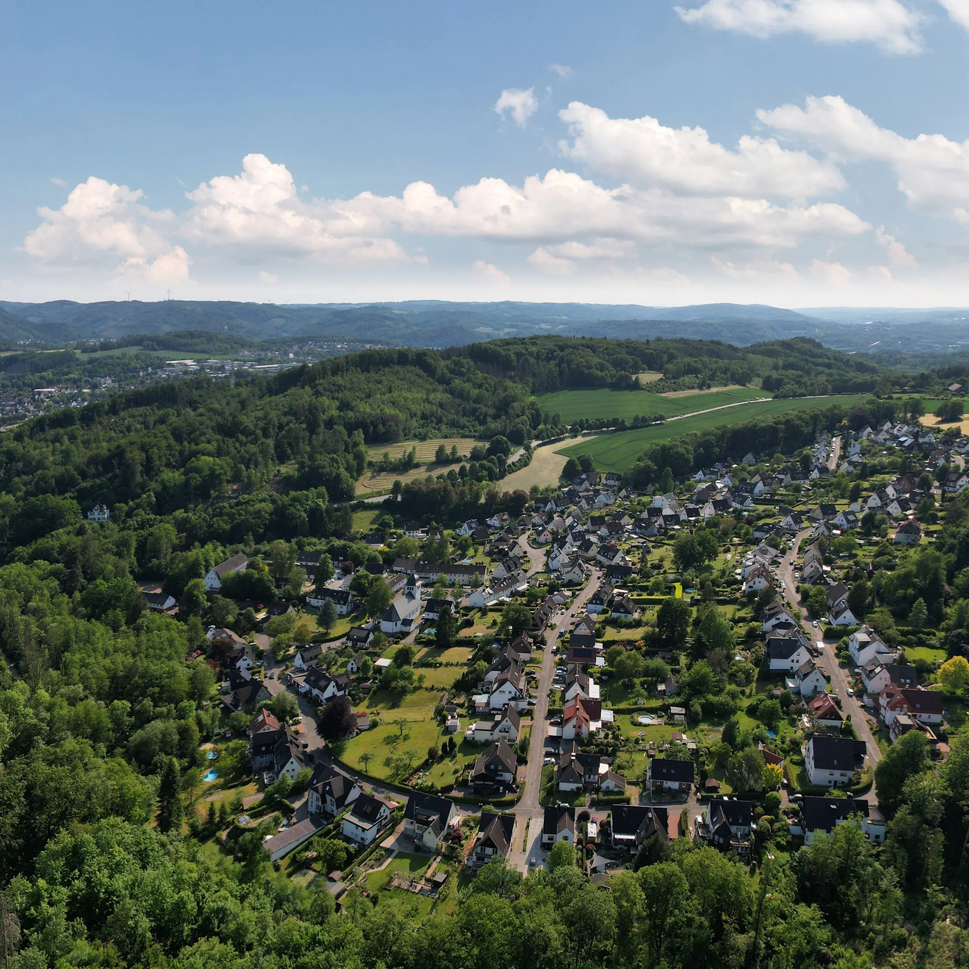 Luftaufnahme einer Wohnsiedlung umgeben von Wäldern und Feldern bei klarem Himmel.