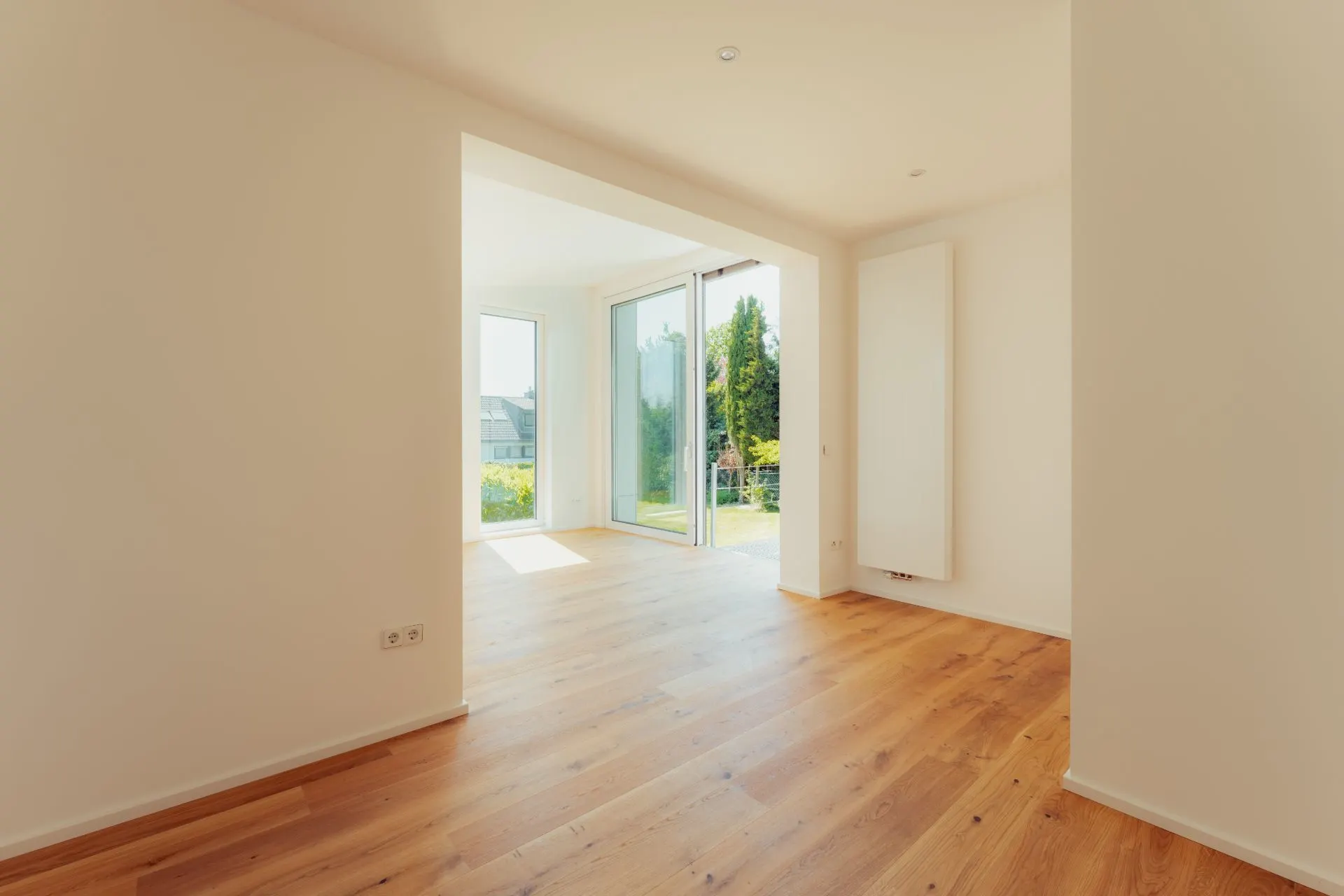 Heller Raum mit Holzboden und großen Fenstern, eine Glastür führt in den Garten.