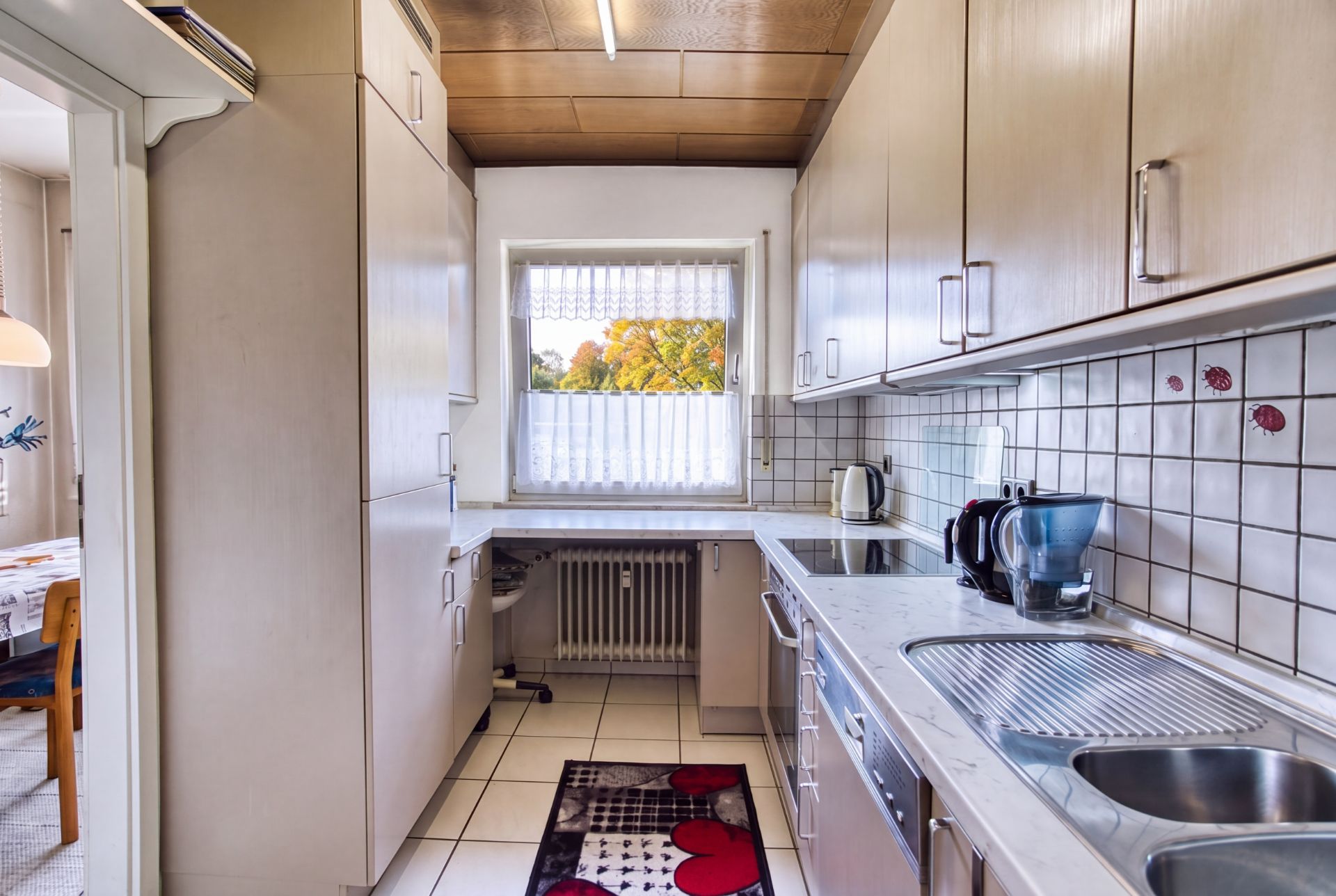 Schmale Küche mit weißen Schränken, Fenster, Spüle, Kaffeemaschine und Fliesenwand.