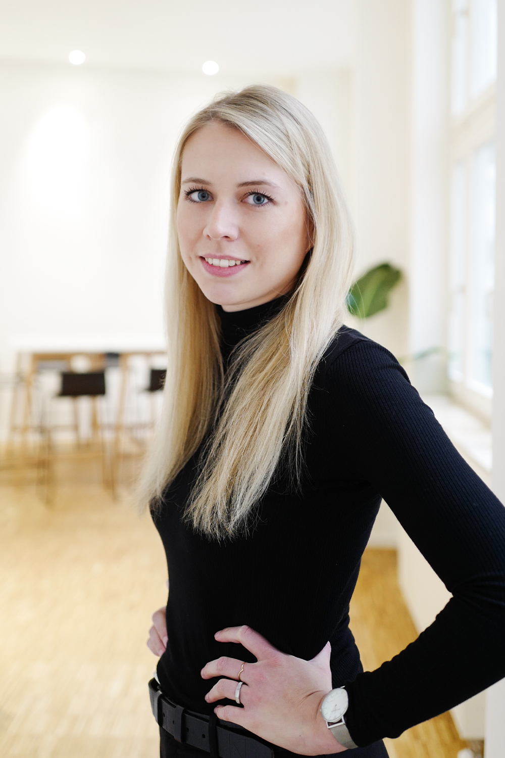 Frau mit langen blonden Haaren steht in einem hellen Raum mit Holzboden und großen Fenstern.