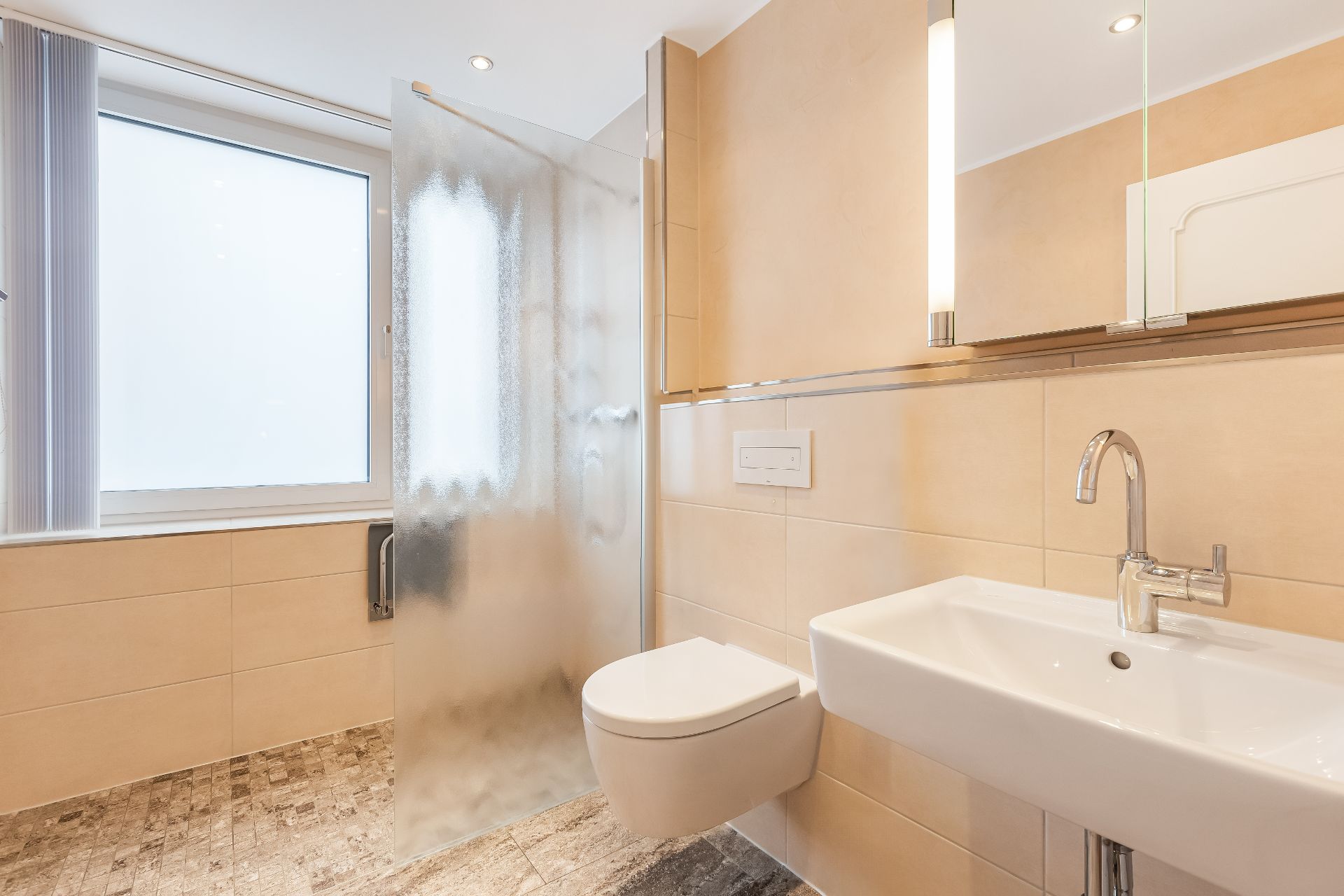 Helles Badezimmer mit Wand-WC, Waschbecken, großer Fensterfront und Duschabtrennung aus Milchglas.