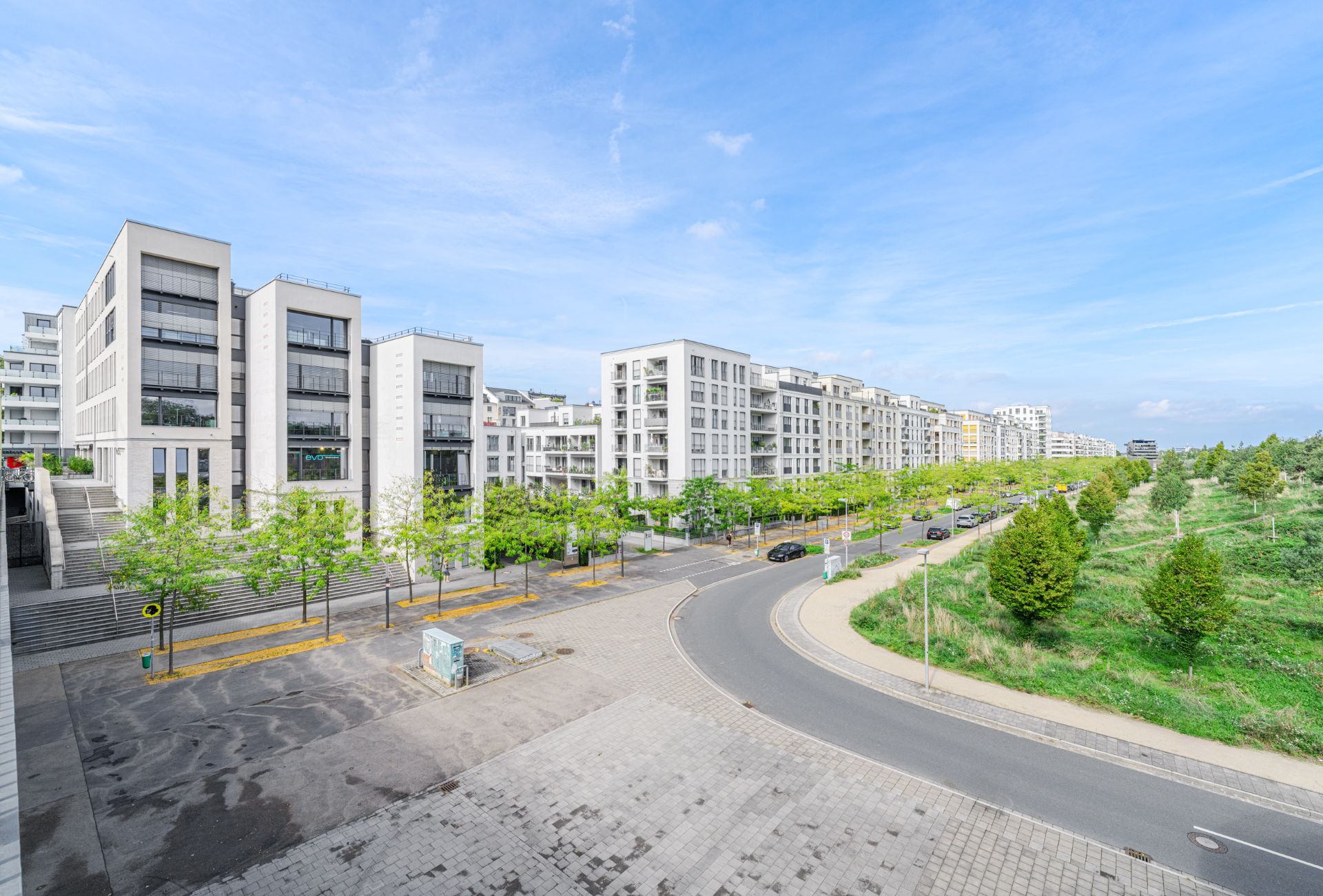 Moderne Wohngebäude mit großen Fenstern und Baumallee an einer Straße bei sonnigem Wetter.