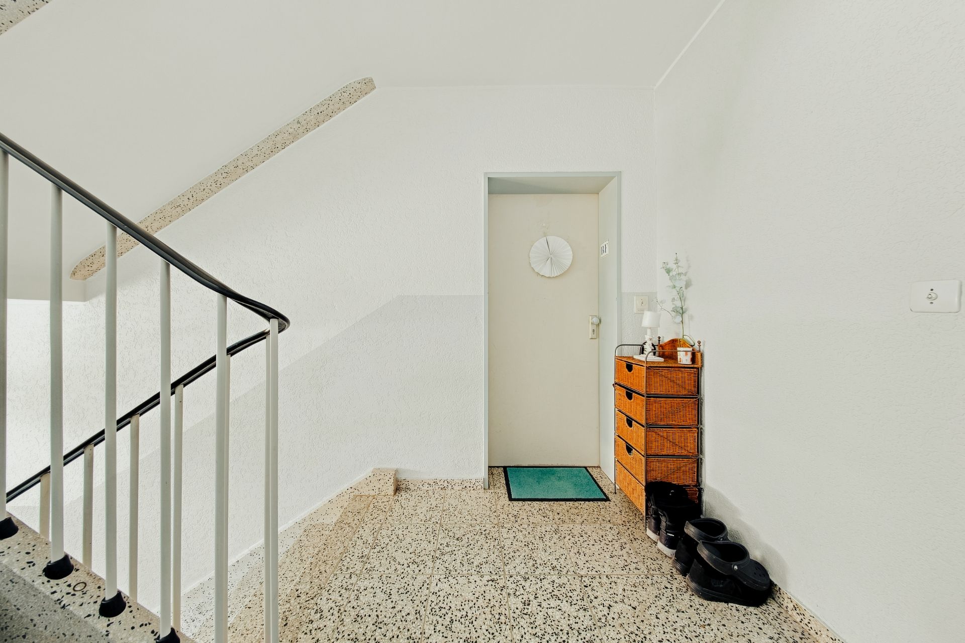 Heller Flur mit Treppe, Tür, Teppich, kleiner Kommode und mehreren Paar Schuhen.
