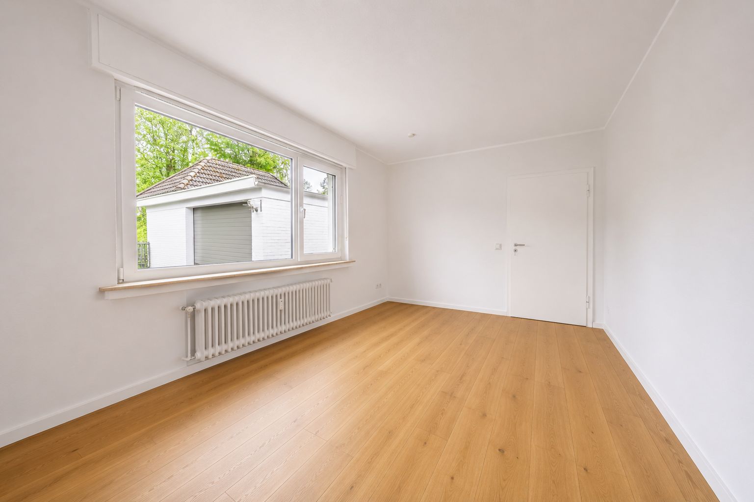 Helles, leeres Zimmer mit großem Fenster, weißer Wand und Holzfußboden.