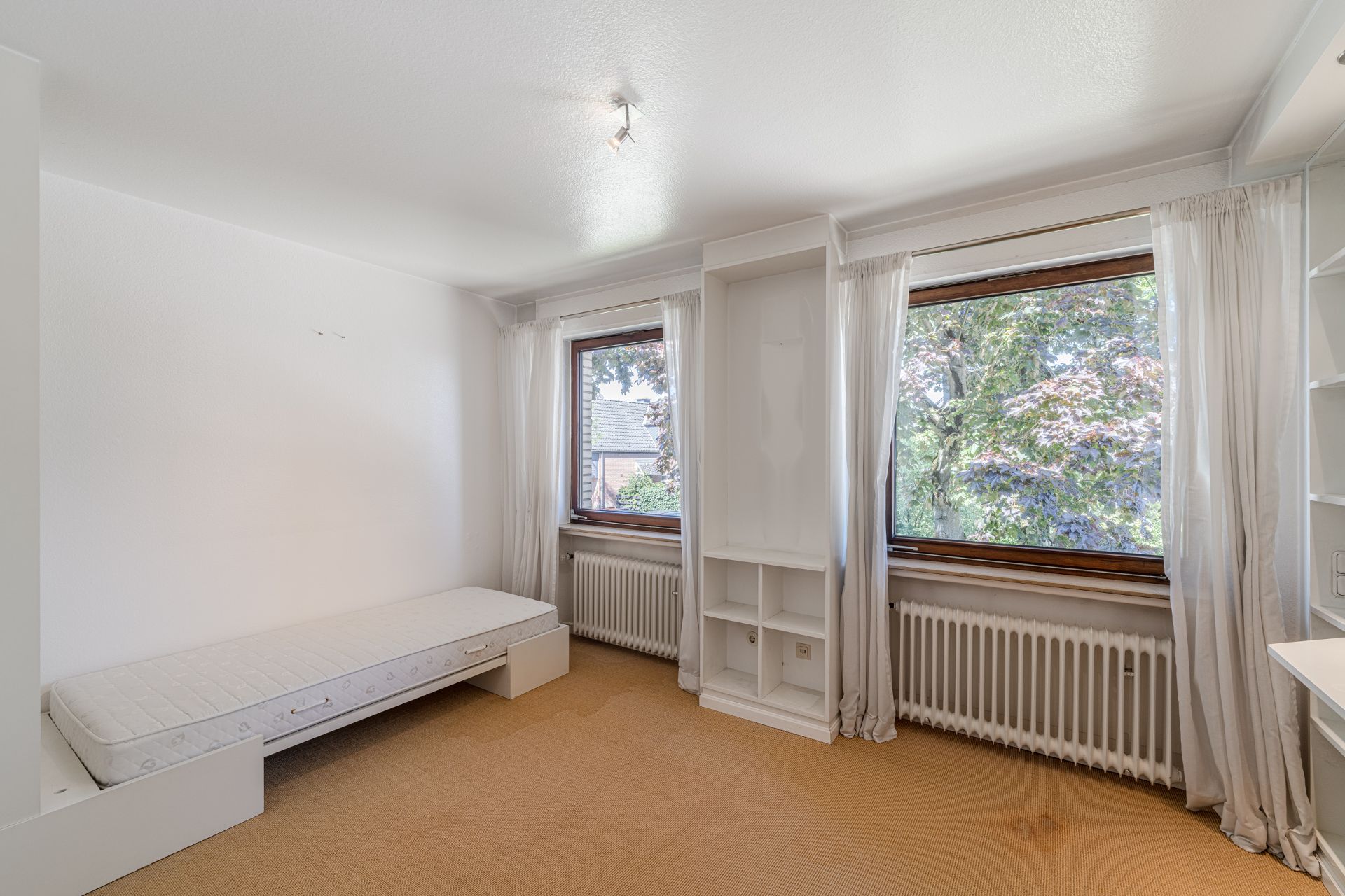 Helles Zimmer mit Einzelbett, Regal, zwei Fenstern mit weißen Vorhängen und Blick auf Bäume.