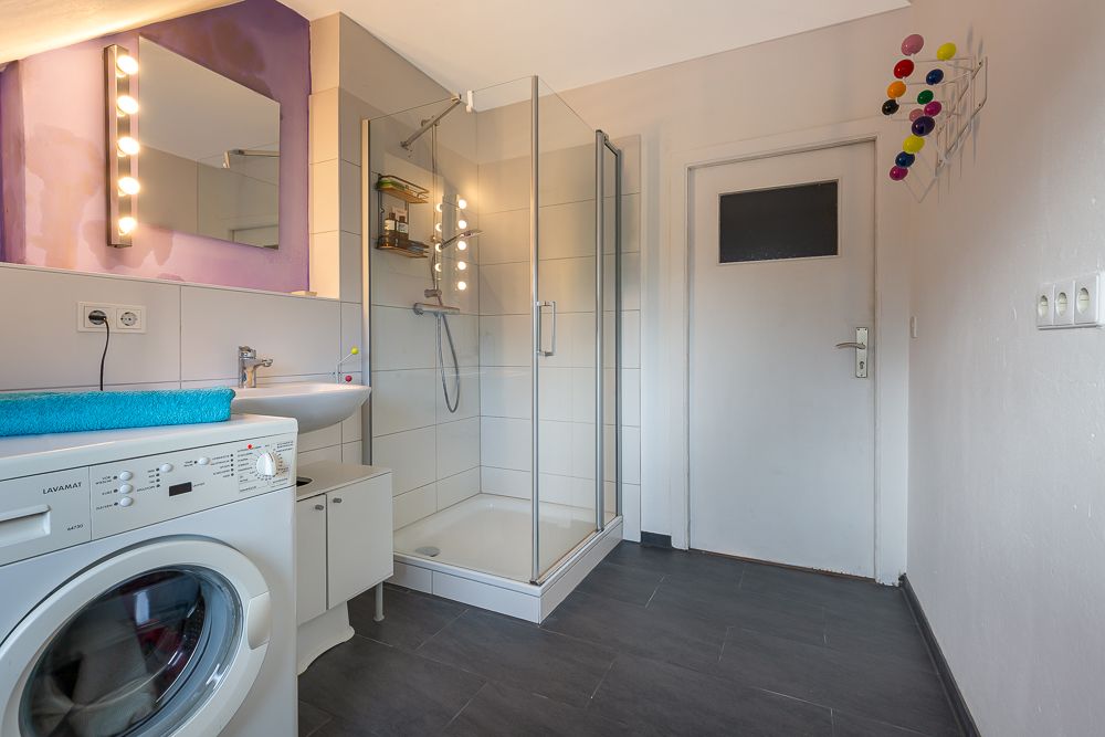 Modernes Badezimmer mit Dusche, Waschmaschine, Waschbecken und beleuchtetem Spiegel.