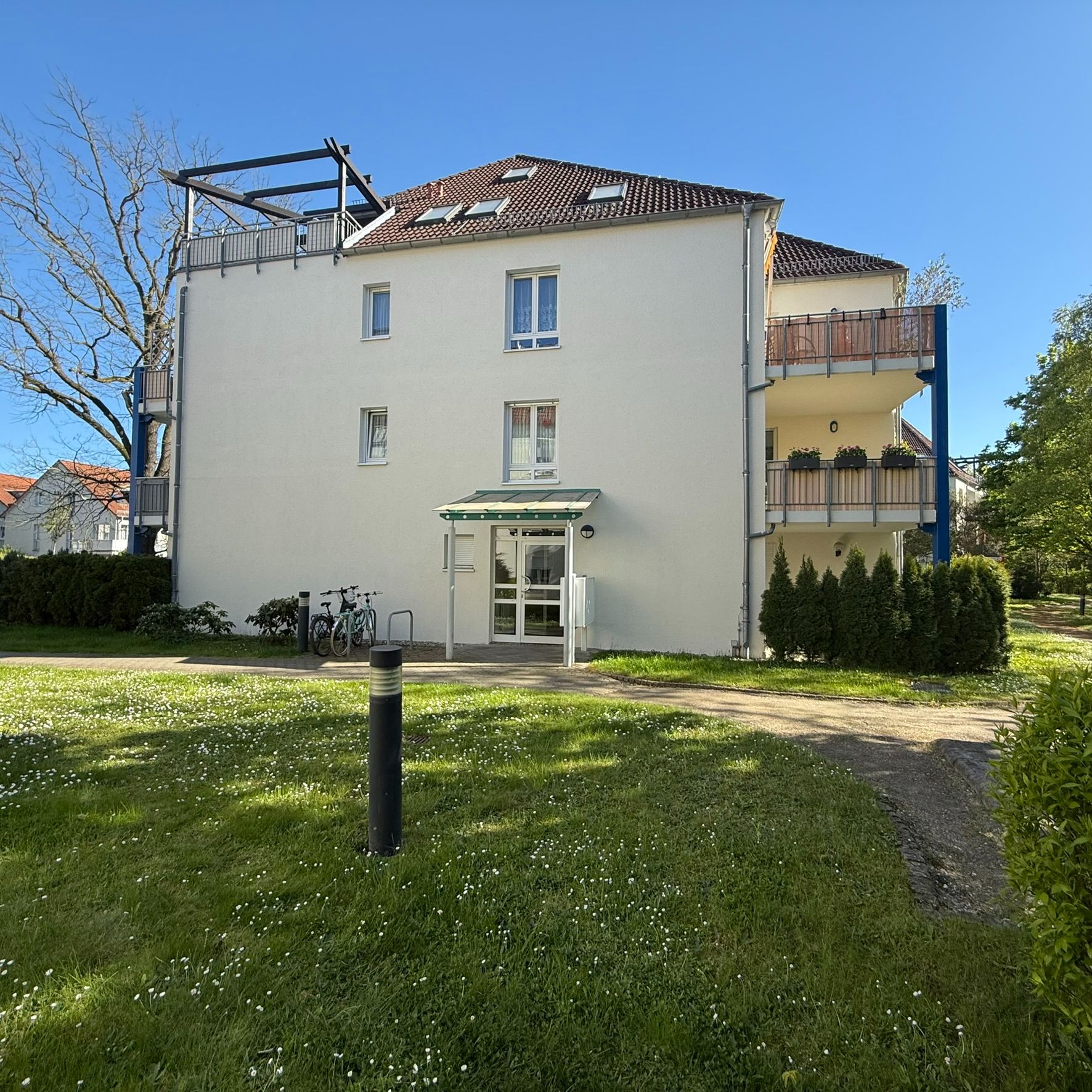 Helles Wohngebäude mit Balkonen, Eingangstür und zwei Fahrrädern vor dem Haus auf einer grünen Wiese.