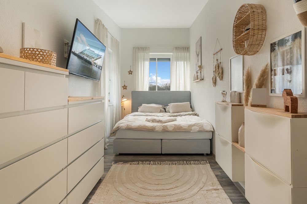 Helles Schlafzimmer mit Doppelbett, weißen Kommoden, Wanddeko und großem Fenster.
