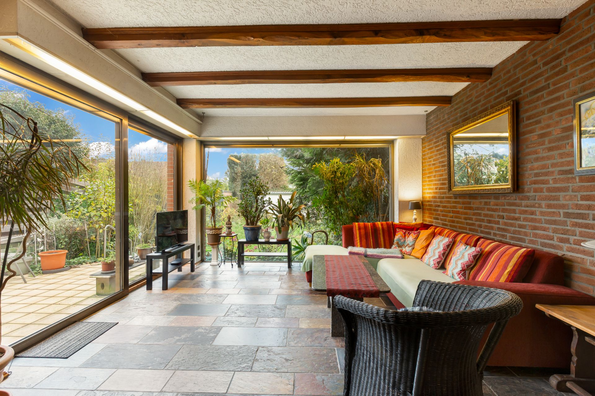 Wohnzimmer mit großen Fenstern, Sofa, Korbstuhl, Pflanzen und Blick in den Garten.