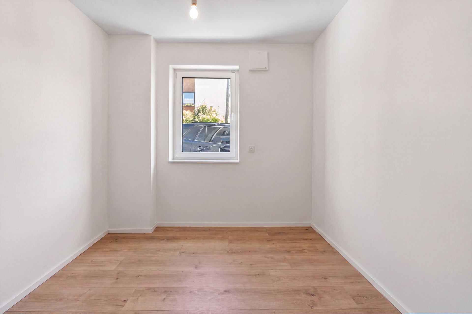 Leerer, schmaler Raum mit Holzfußboden, weiß gestrichenen Wänden und einem Fenster an der Stirnseite.