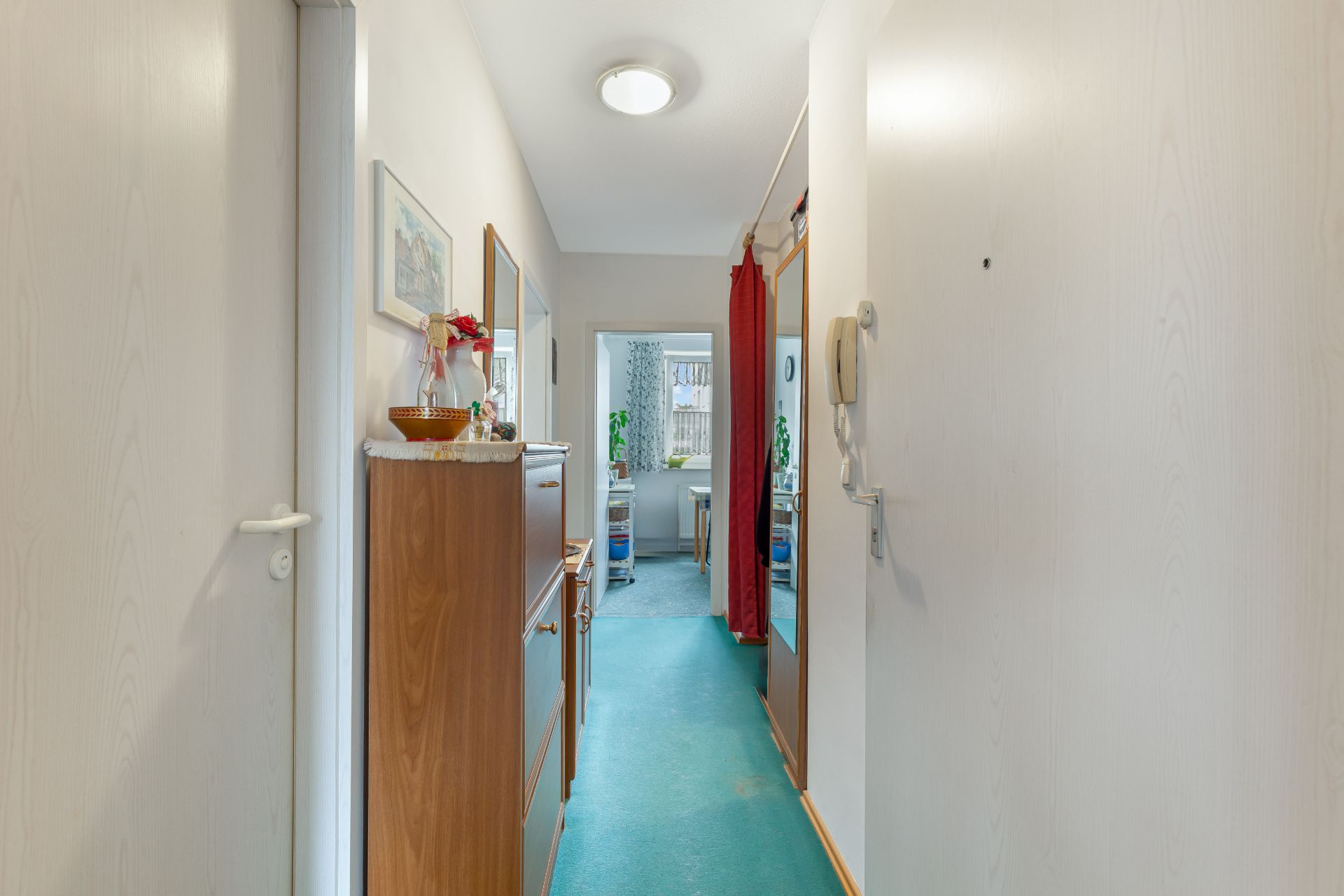 Heller Flur mit grünem Teppich, Spiegel, Kommode und Blick in ein weiteres Zimmer.
