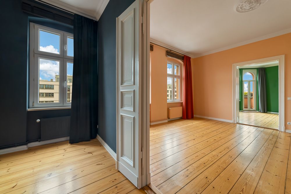Blick in drei helle Zimmer mit Holzboden, bunten Wänden und großen Fenstern mit Vorhängen.
