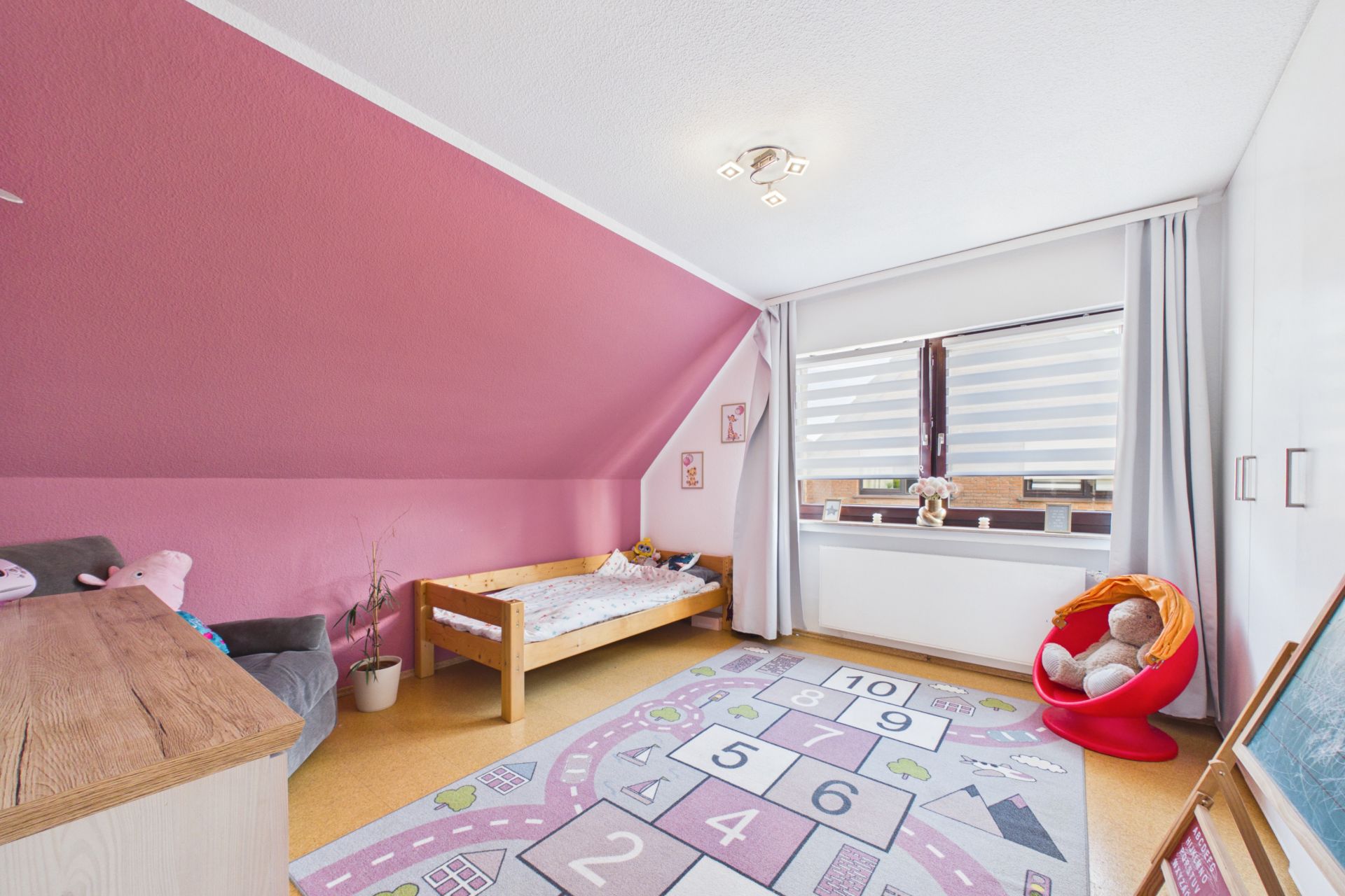 Helles Kinderzimmer mit rosa Wand, Einzelbett, Spielteppich und großem Fenster mit Vorhängen.