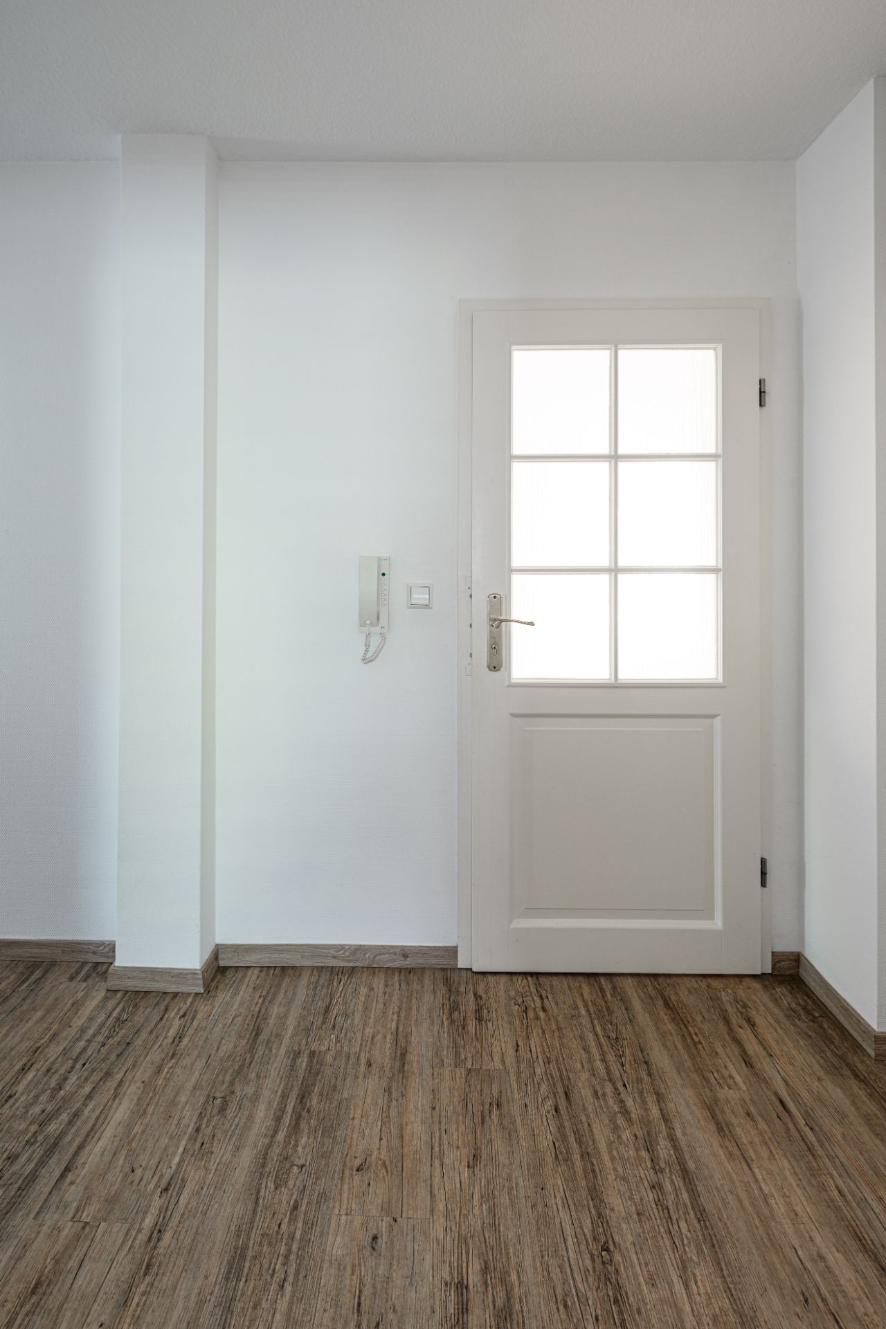 Weiße Tür mit Glaseinsatz und Gegensprechanlage an einer weißen Wand, Holzboden.