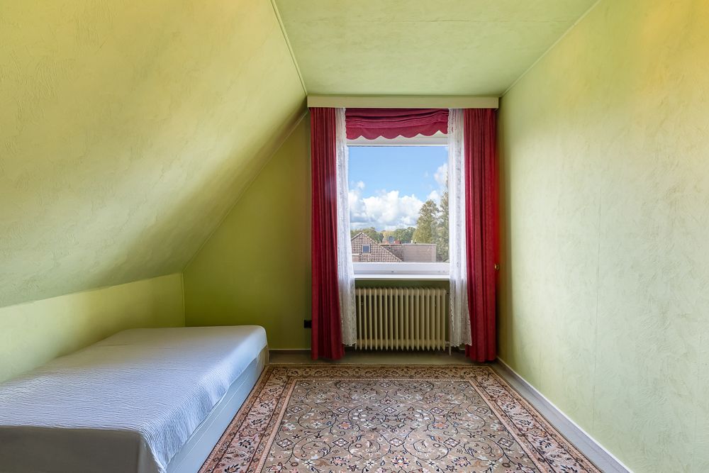 Kleines Zimmer mit Einzelbett, Teppich und Fenster mit roten Vorhängen.