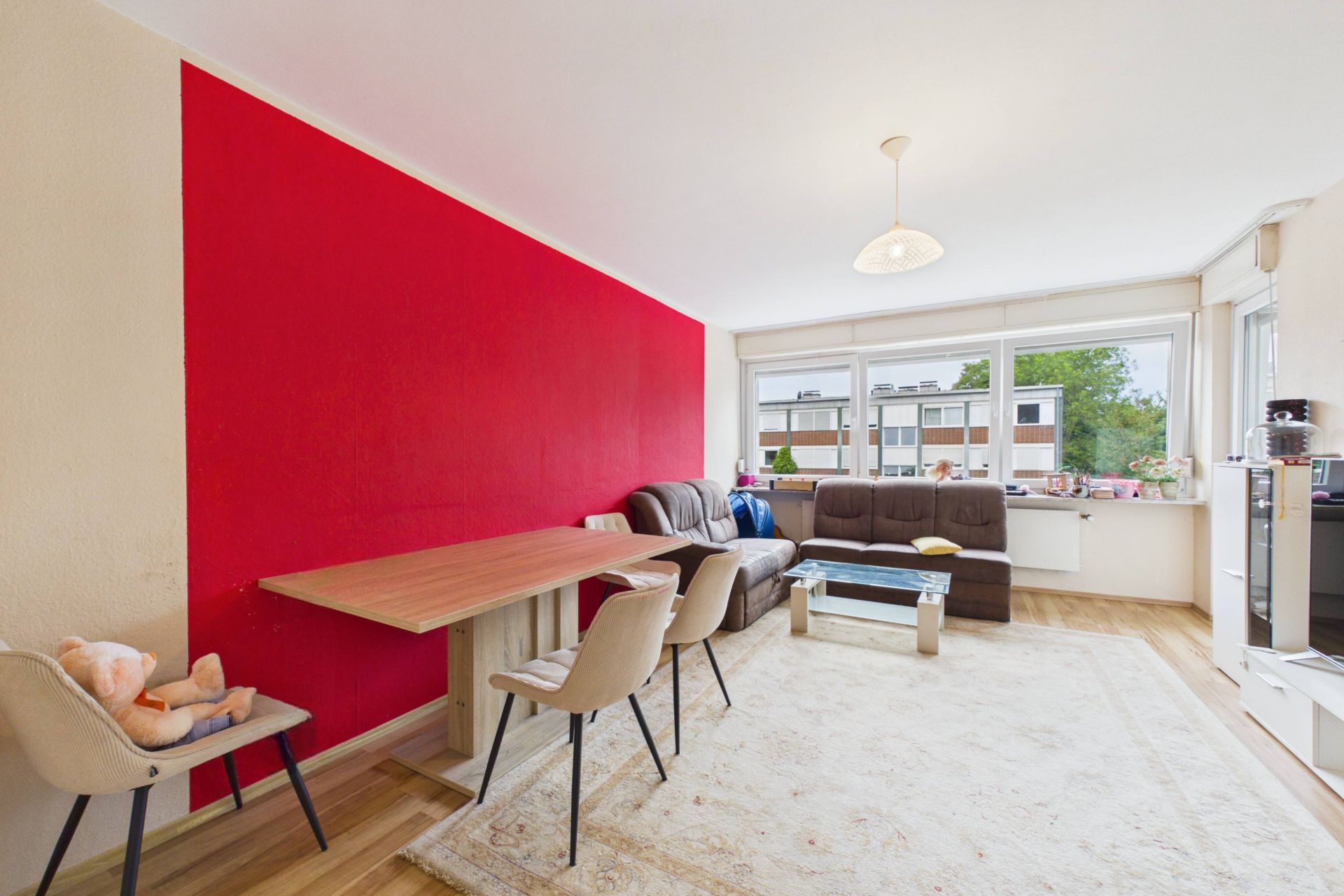 Helles Wohnzimmer mit roter Wand, Essbereich, Sofa und großem Fenster mit Blick nach draußen.