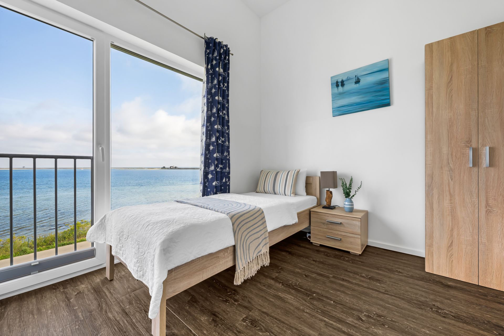 Helles Schlafzimmer mit Einzelbett, Kleiderschrank und Meerblick durch großes Fenster.