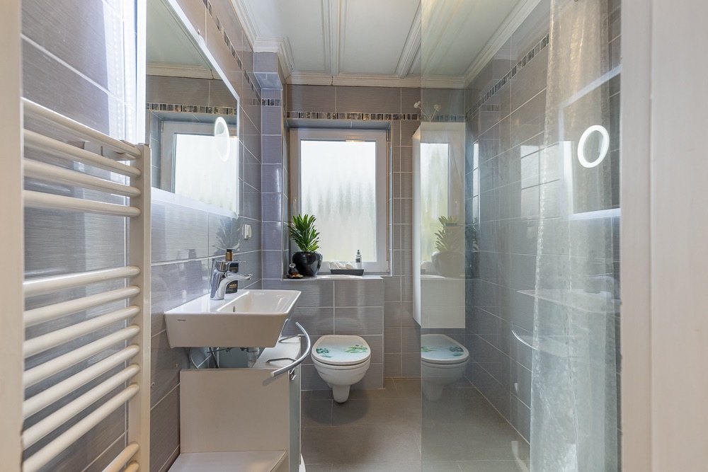 Modernes Badezimmer mit Waschbecken, Toilette, Dusche und zwei Fenstern mit Milchglas.
