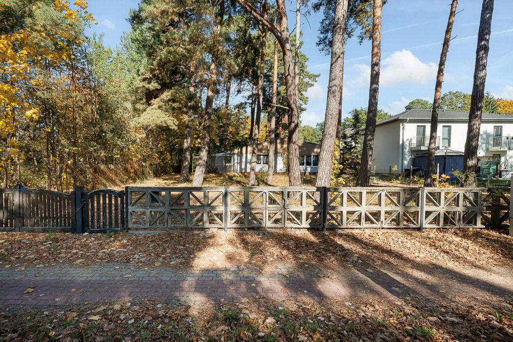 Holzzaun vor einem Haus mit Bäumen und Herbstlaub am Boden.