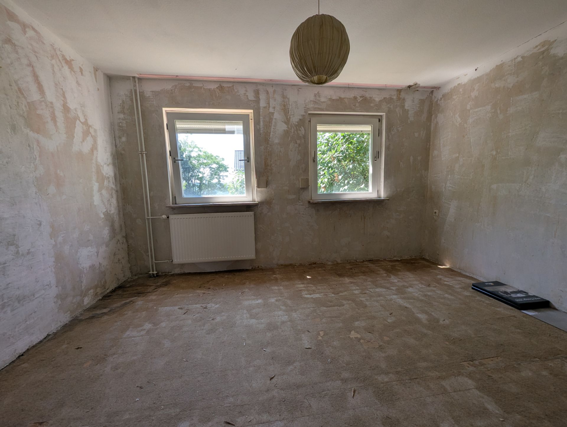 Leeres Zimmer mit zwei Fenstern, abblätternden Wänden und einer Deckenlampe.