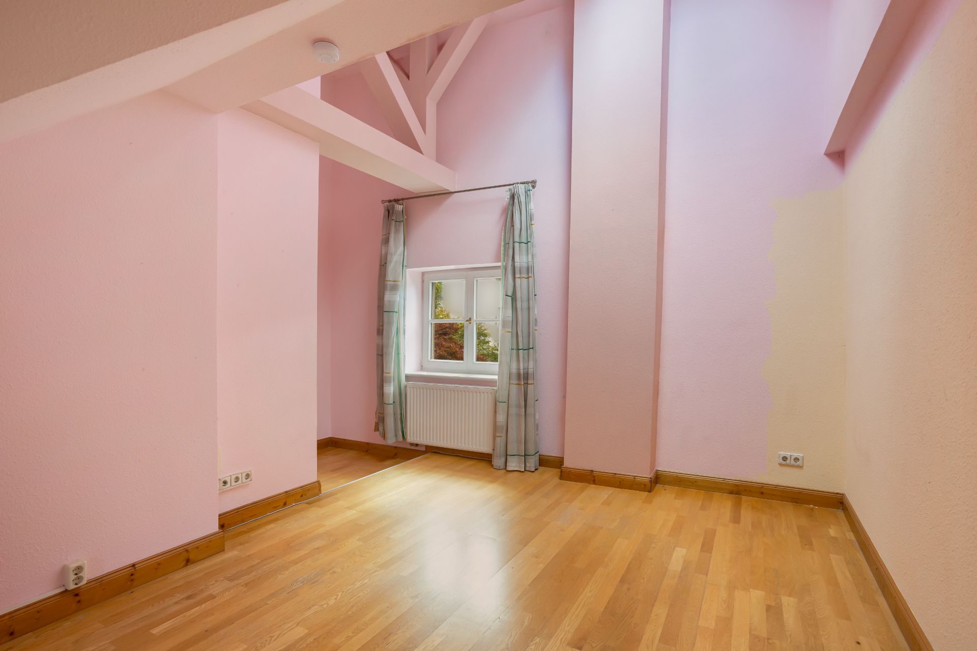 Leerstehendes Zimmer mit rosa Wänden, Holzfußboden und kleinem Fenster mit Vorhängen.