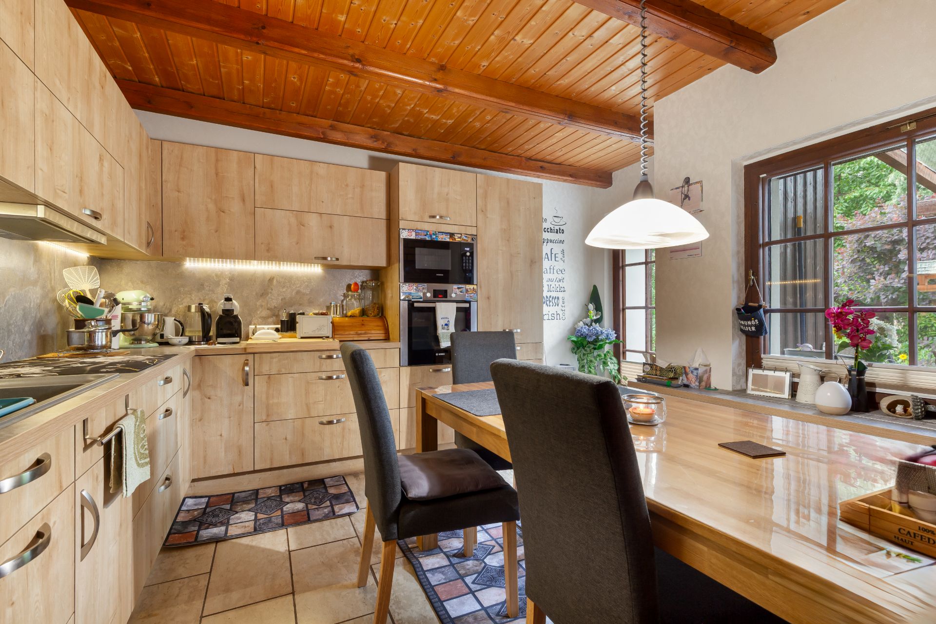 Moderne Küche mit Holzmöbeln, Esstisch, Stühlen und großem Fenster mit Aussicht in den Garten.