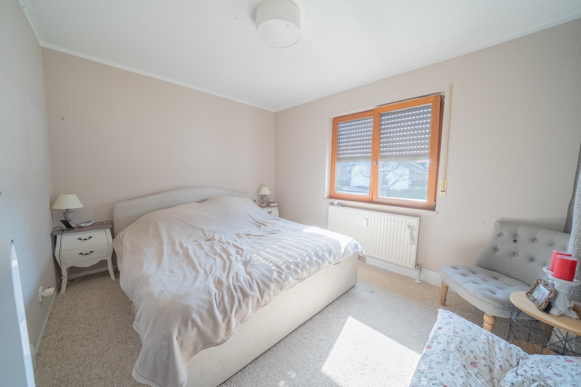 Helles Schlafzimmer mit Doppelbett, zwei Nachttischen, Fenster und zwei Stühlen.