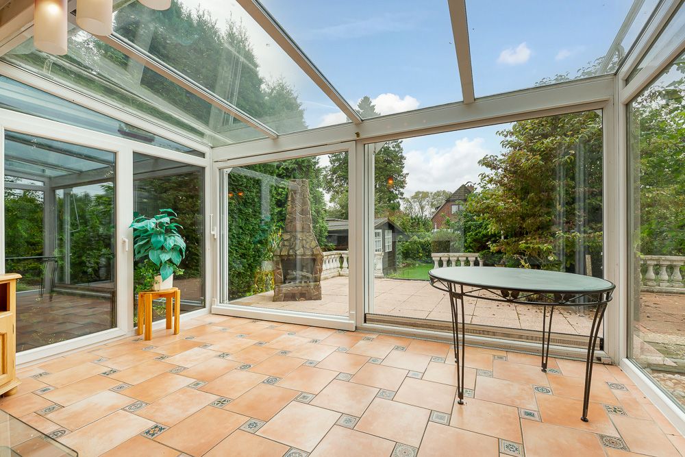 Wintergarten mit Glasdach, Fliesenboden, kleinem Tisch und Blick in den Garten.