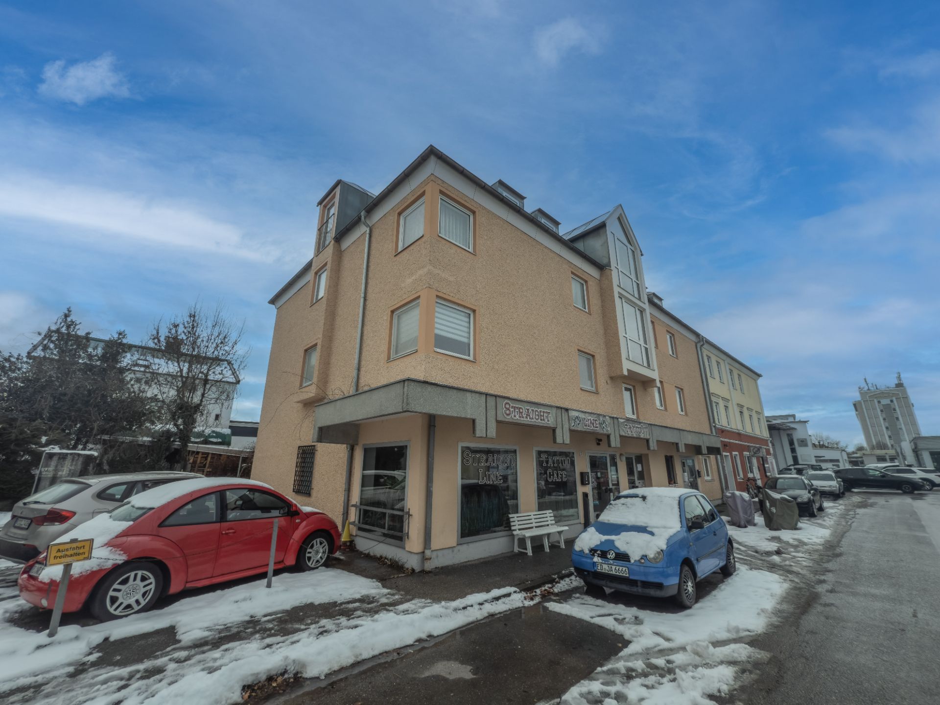 Mehrstöckiges Wohnhaus mit geparkten Autos und Schnee am Straßenrand.