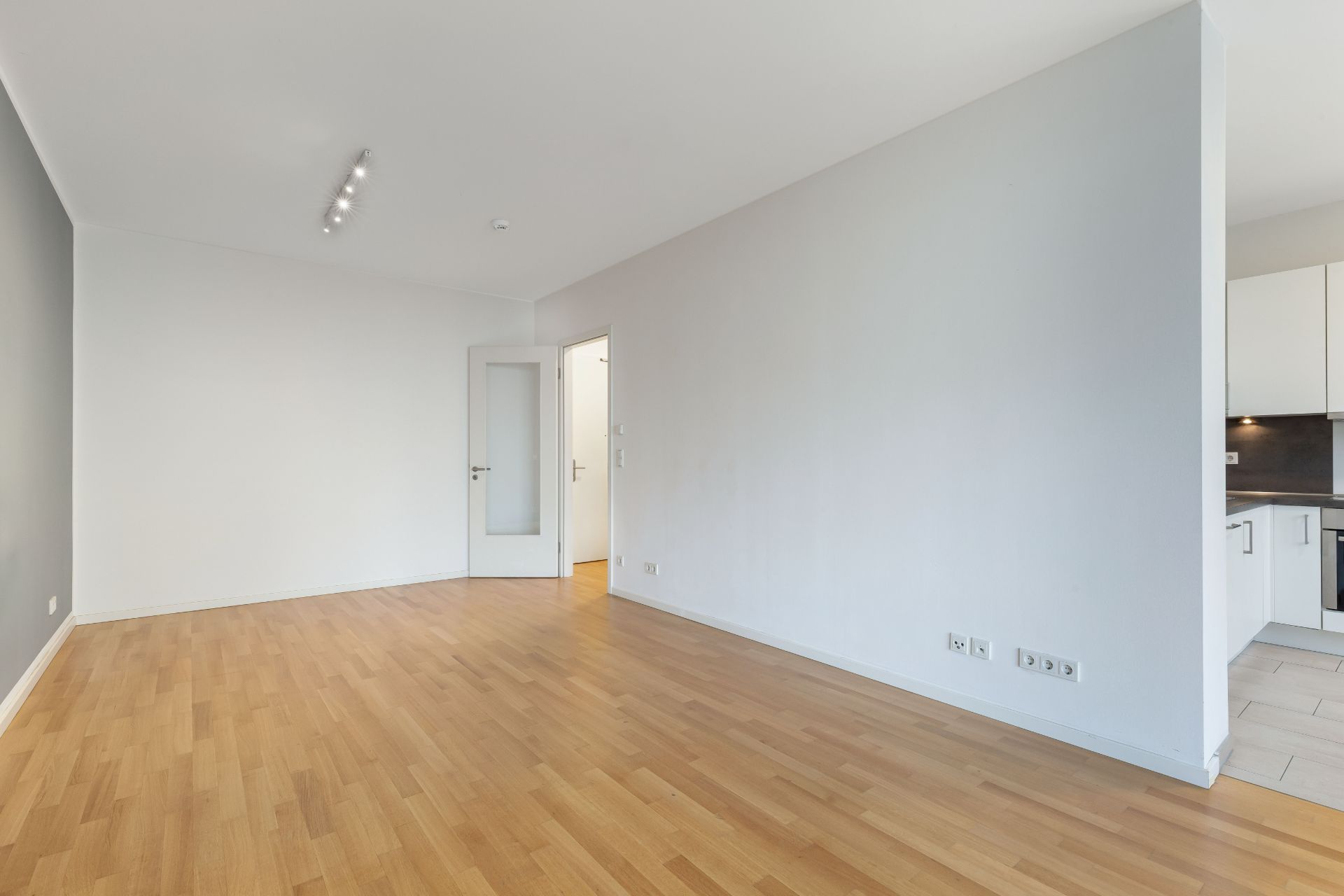 Helles, leeres Wohnzimmer mit Holzfußboden, Tür und Blick in eine weiße Einbauküche.