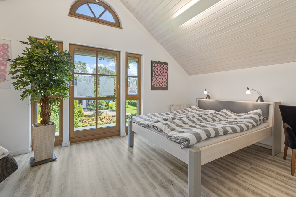 Helles Schlafzimmer mit großem Fenster, Doppelbett, Stehlampe und Zimmerpflanze.