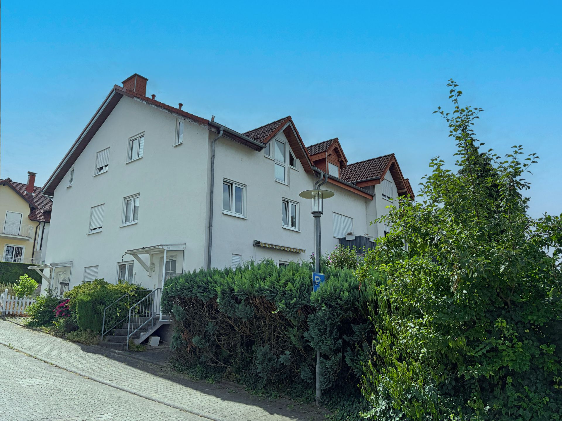 Weißes Wohnhaus mit rotem Dach, Treppe zum Eingang, umgeben von Sträuchern und Bäumen.