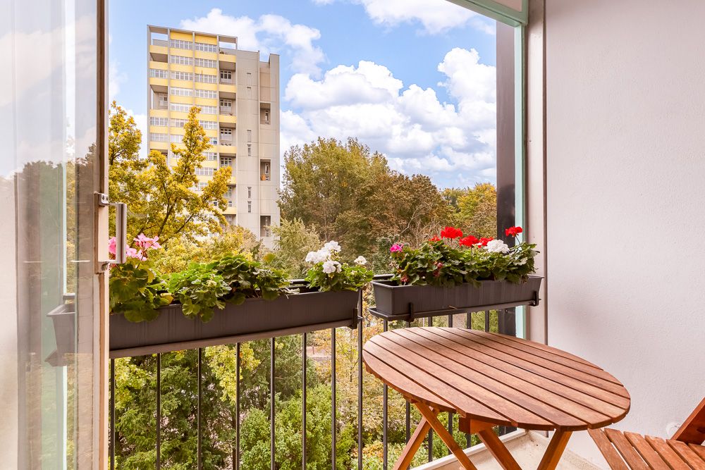 Kleiner Balkon mit Holztisch, Blumen auf dem Geländer und Blick auf Bäume und ein Hochhaus.